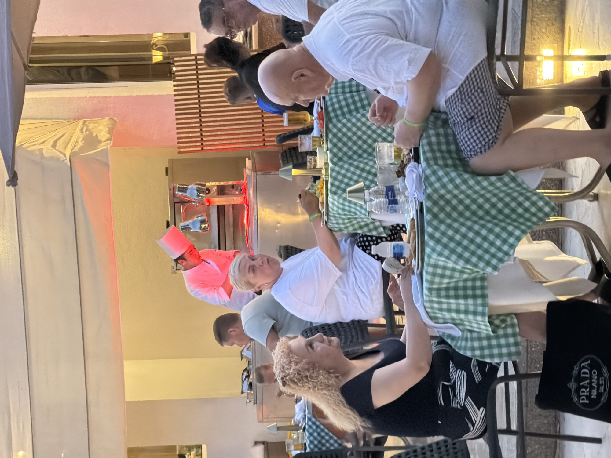 The image depicts an outdoor dining scene at a restaurant, possibly located in Sharm El Sheikh, Egypt, given the warm lighting and a glimpse of Mediterranean-style architecture. In the foreground, two individuals are seated at a table covered with a green and white checkered tablecloth. The woman on the left has curly blonde hair and is wearing a black top with a patterned skirt. The man on the right is bald, wearing a white polo shirt and checkered shorts. Both appear to be engaged in conversation and are interacting with food and drinks on the table. Behind them, a chef in a white uniform and hat is visible near a food preparation area with a large oven or grill. Other patrons are seated at tables in the background, and the overall atmosphere suggests a lively evening meal. The lighting is warm and somewhat dim, with a red glow emanating from the area behind the chef, likely from heat lamps or decorative lighting.