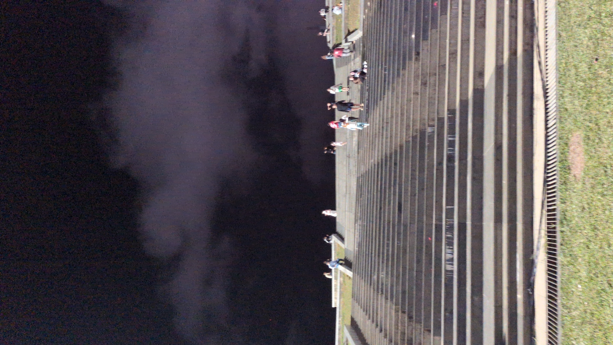 The image depicts a wide, tiered staircase at night, illuminated by ambient lighting. Several individuals are scattered across the steps, some walking and others sitting. The sky above is dark, with visible clouds, suggesting an overcast or possibly stormy night. A grassy area with a drainage grate is visible at the bottom of the frame, leading up to the stone steps. This staircase is located at the Lam Vien Square in Da Lat, Vietnam.