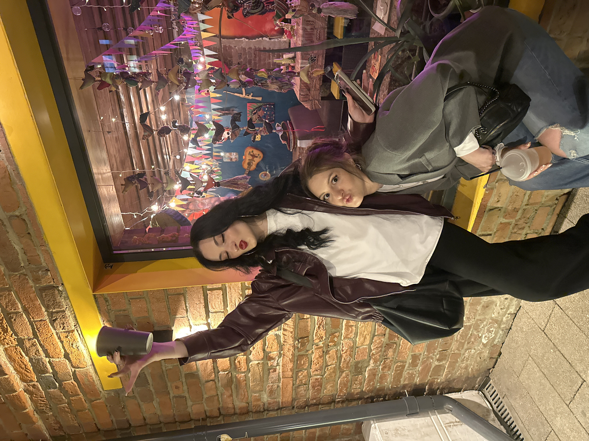 Two women are posing for a photo in an indoor location, likely a restaurant or bar. One woman is wearing a white t-shirt, a burgundy leather jacket, and black pants. She is holding a gray cup in her hand. The other woman is wearing a gray blazer, a white shirt, and ripped jeans. She is holding a phone in one hand and a plastic cup in the other. A black purse is hanging from her shoulder. The background features a brick wall, a decorative window frame, and colorful decorations, including string lights, tassels, and fabric cutouts. Based on the available information, the location could be a bar or restaurant in Krasnoyarsk, Russia.
