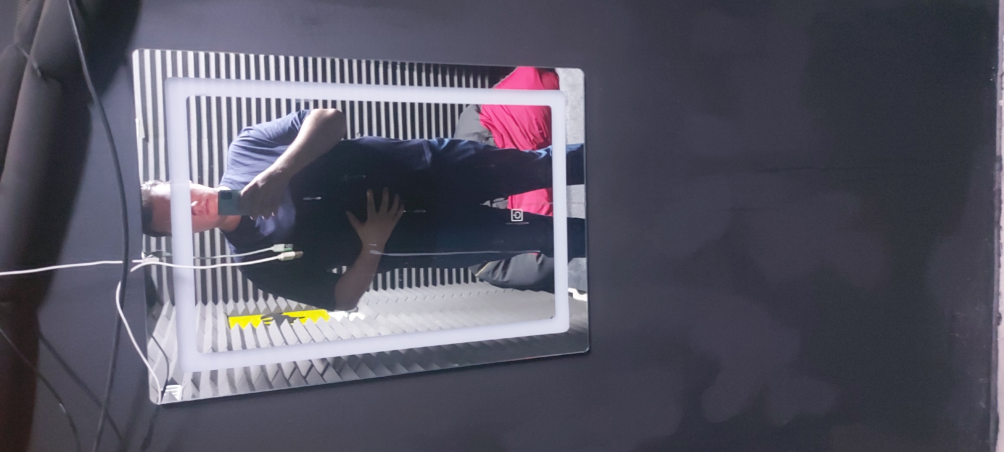 A man in a navy blue shirt and navy blue pants is taking a picture of his reflection in a rectangular mirror with a white light frame. The mirror is mounted on a dark gray wall with what looks like padded insulation to the left. The background reflected in the mirror consists of black and white vertical stripes, possibly a wall covering or fabric. There is a pink garment or cloth visible above the man's reflected torso. Cords are plugged into the bottom of the mirror frame. The image was taken in Tashkent, Uzbekistan.