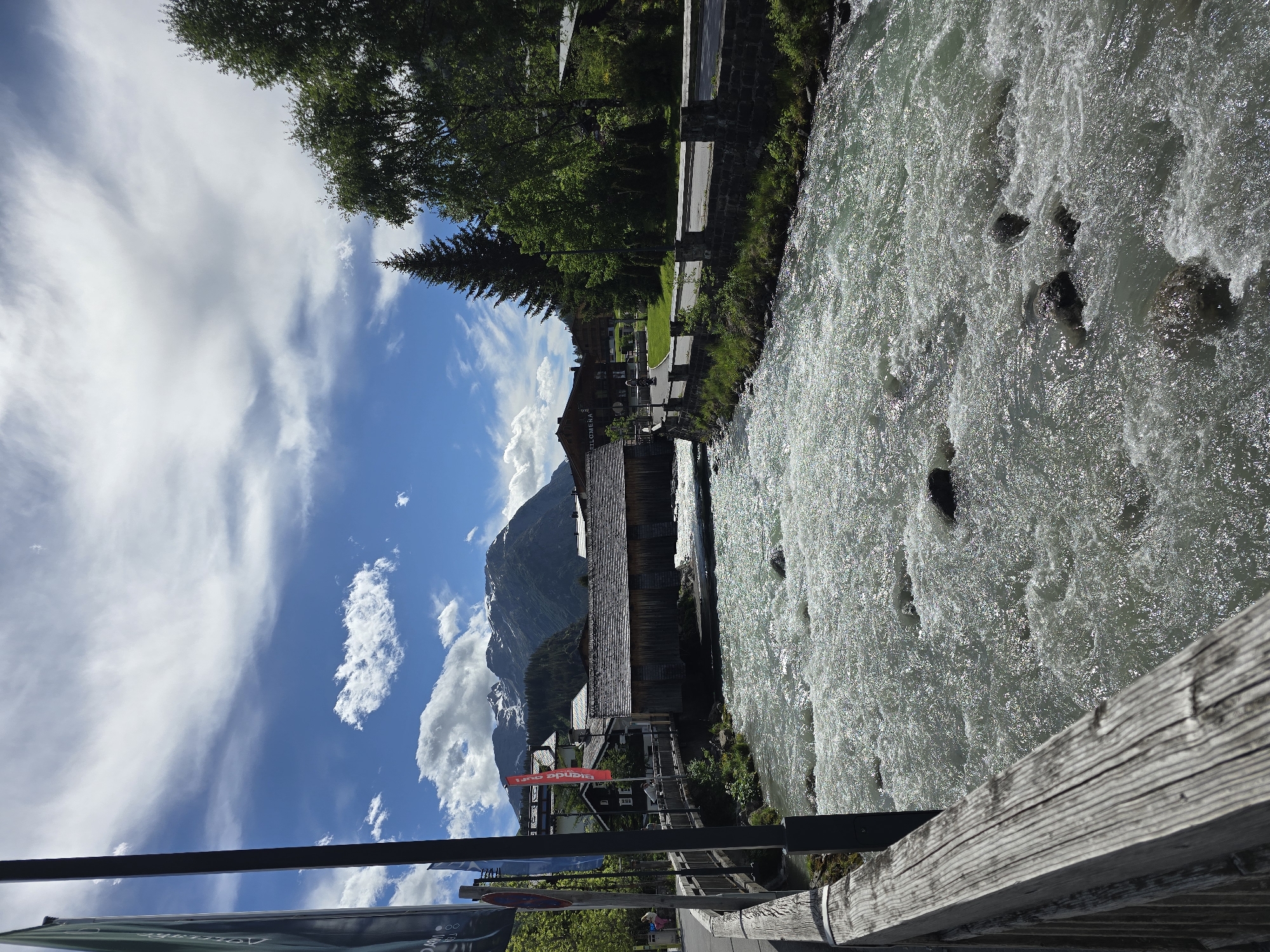 The image depicts a fast-flowing, rocky river in Lech, Austria, with a wooden bridge and buildings in the background. The river's surface is heavily textured with ripples and white water, suggesting a strong current. On the left bank of the river, a wooden railing runs along the foreground, leading to a dark pole. Flags are visible on the left side of the image, with text that is partially obscured. A road with a stone wall runs along the right bank, with trees and more buildings situated behind it. In the distance, a snow-capped mountain range looms under a partly cloudy sky. The overall scene is a picturesque alpine landscape, capturing the natural beauty of Lech.