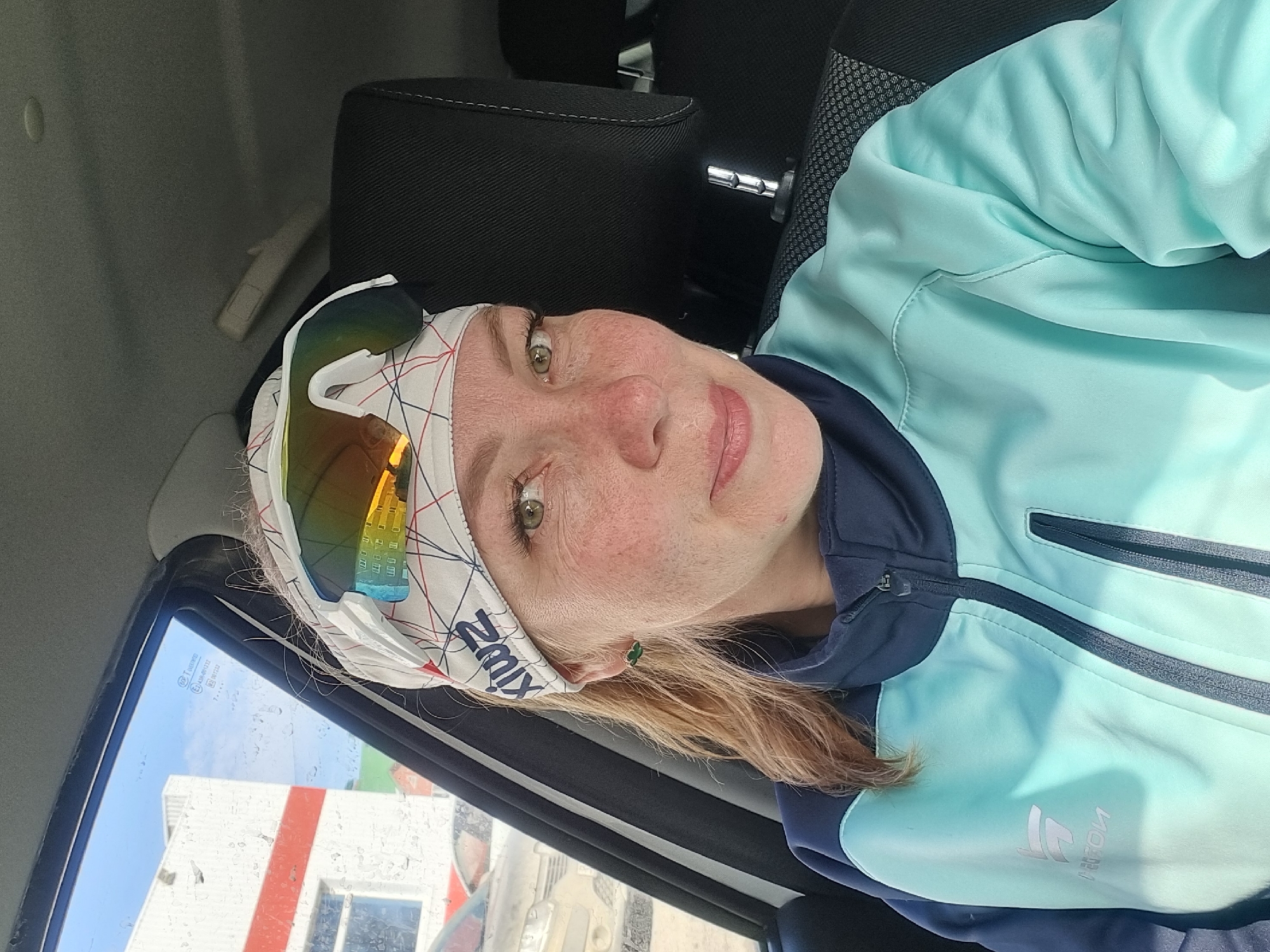 The image depicts an adult female positioned inside a vehicle, likely in the passenger seat, looking forward towards the camera. She has fair skin with visible freckles across her nose and cheeks, green eyes, and light brown hair pulled back. She is wearing a light blue and dark blue zippered athletic jacket with a small logo on the left chest, and a white head bandana with a red and blue grid pattern and a dark blue "Swix" logo. Mirrored sports sunglasses are resting on top of the bandana. Small green earrings are visible on her earlobes. The vehicle's interior features a dark fabric headrest and a gray ceiling. Through the vehicle's window, which is speckled with water droplets, an outdoor scene is visible. This includes a large white building with a prominent horizontal red stripe and multiple windows, indicative of a commercial structure. Above the building, a light blue sky with scattered clouds is present. Other vehicles are partially visible in the foreground outside. Reflections in the sunglasses show a multi-story building with yellow or beige walls and dark windows. The exact location in Chaikovsky, Russia, cannot be precisely determined from the visible background elements, but the scene suggests a commercial or urban area.