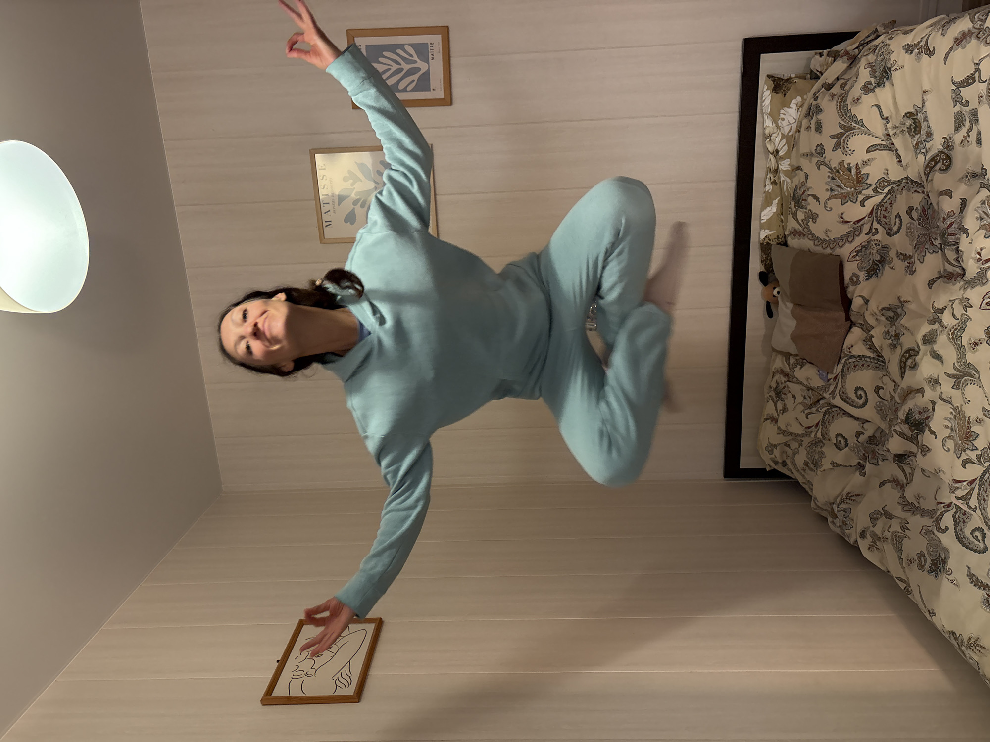 A woman wearing a blue sweatsuit is levitating in a room. The room has light-colored walls with a vertical striped pattern. There are three framed art prints on the wall behind her and one on the floor in front of her. A large bed covered in a patterned comforter is in the right side of the frame. A lamp is in the upper left corner. The location is an indoor space in an unknown city, but not Gelendzhik, Russia.