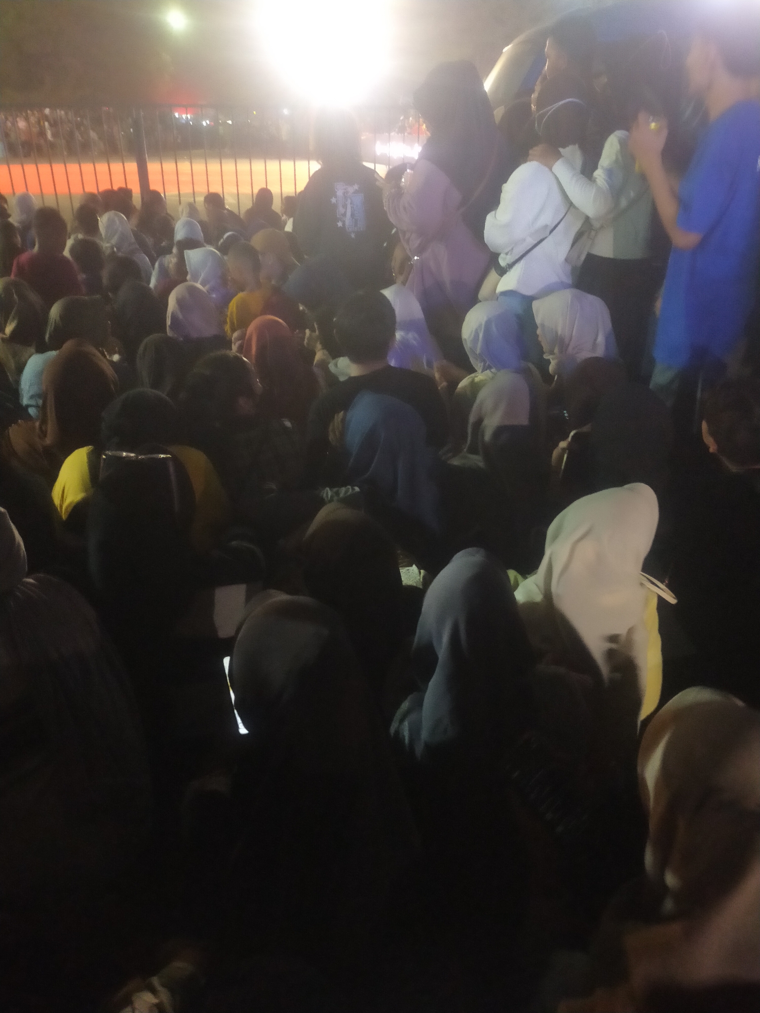 The image depicts a nighttime scene of a crowded gathering in Slawi, Indonesia, under bright artificial lighting. A dense mass of people, mostly young adults, are closely packed together. Many are wearing headscarves or hoods, with a variety of clothing colors including dark tones, white, blue, and yellow. Some individuals are interacting, with one person in the center appearing to embrace another from behind. In the background, a fence with orange horizontal elements is visible, and beyond that, indistinct structures and further crowds are suggested by lights and silhouettes. The overall impression is of a lively outdoor event or assembly.
