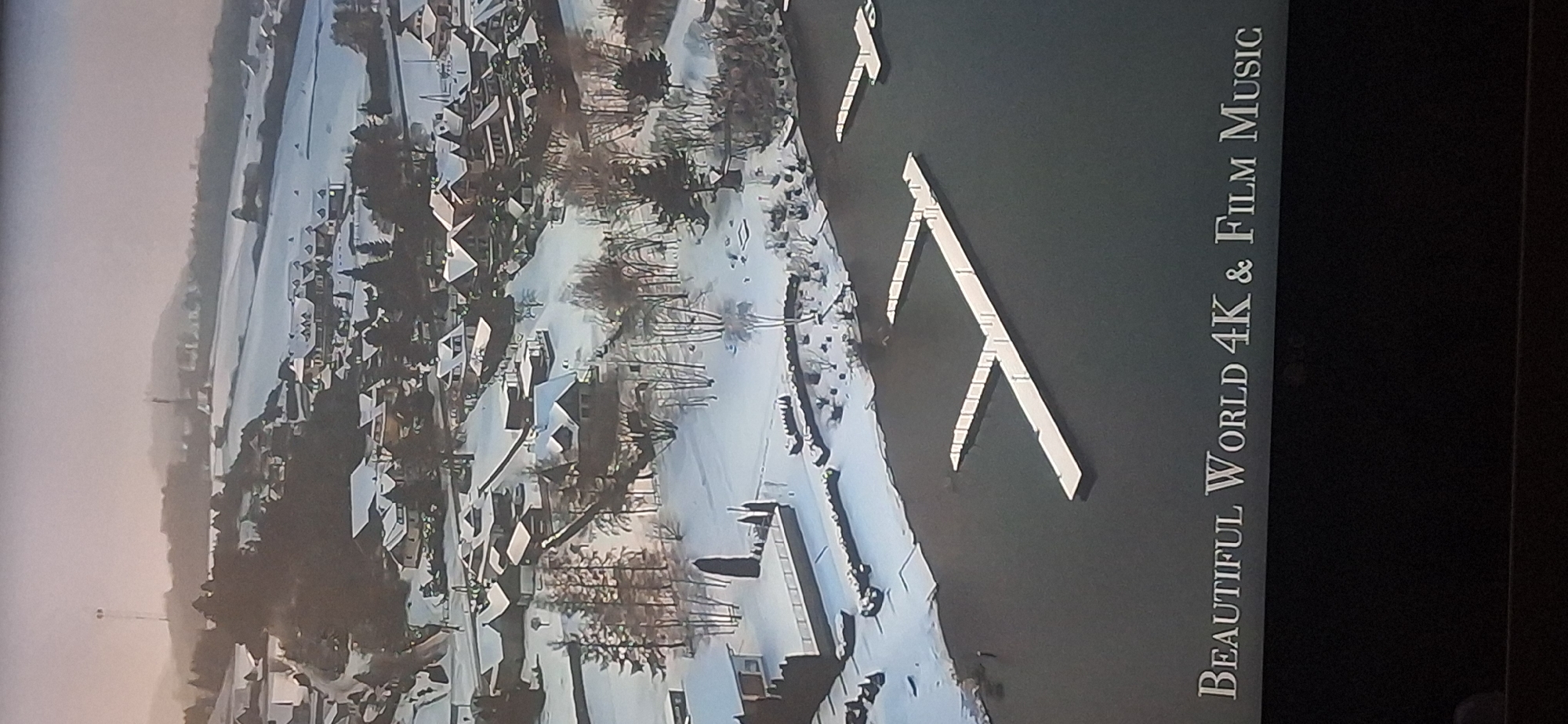 The image shows an aerial view of a snow-covered landscape with several buildings nestled amongst trees. The buildings appear to be houses with pitched roofs, and some have lights on, suggesting occupancy. There is a body of water in the foreground, likely a lake or river, with two modern-looking white piers extending into it. The landscape is hilly and appears to be a rural or semi-rural setting. In the distance, more hills are visible, also covered in snow, with some structures that could be further buildings or communication towers. The sky is overcast, creating a soft, diffused light. The bottom of the image displays text that reads "BEAUTIFUL WORLD 4K & FILM MUSIC". There is no discernible evidence or context to associate this scene with the city of Bila Tserkva, Ukraine.