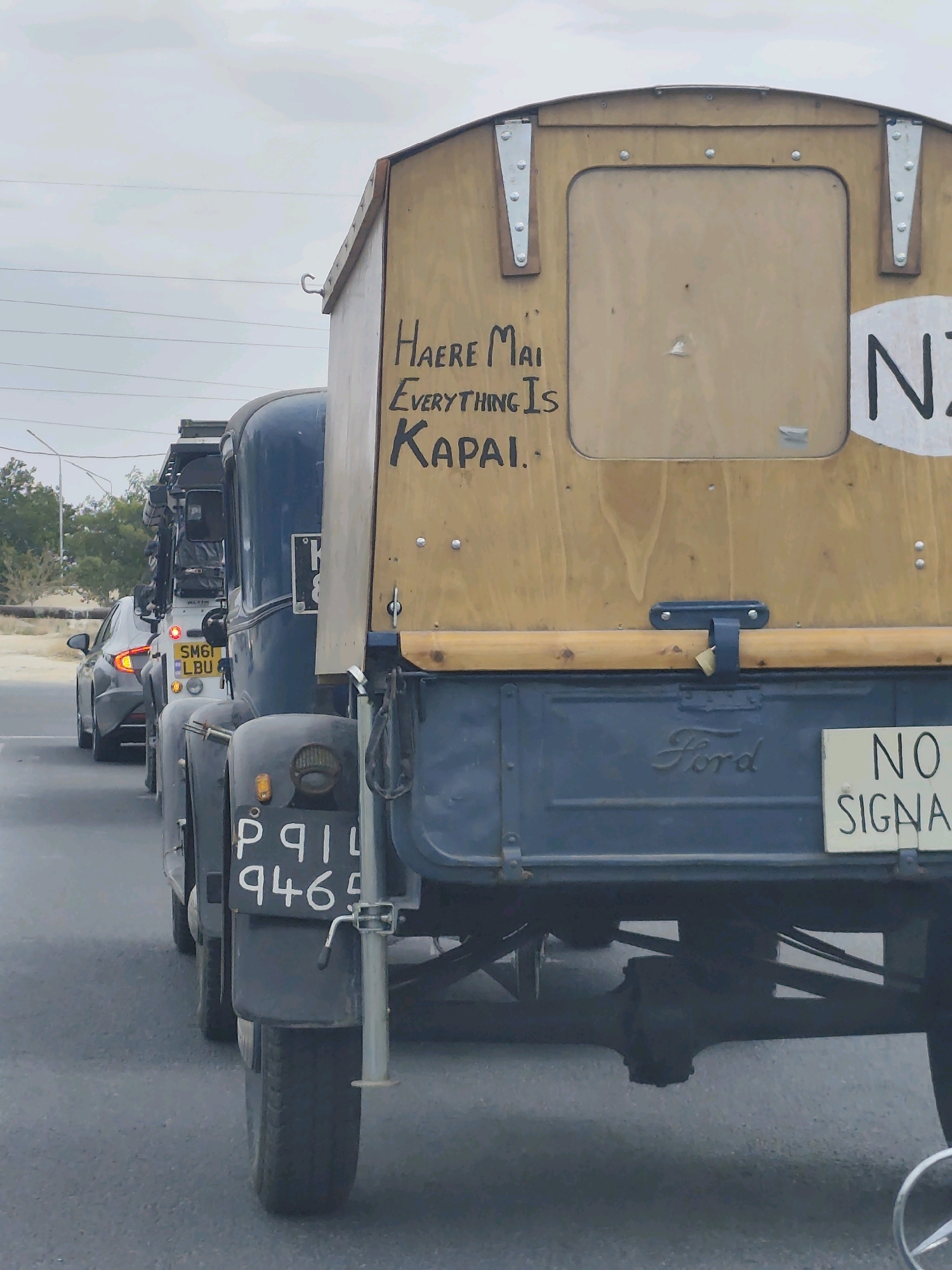 The image shows the rear of a vintage blue Ford truck with a wooden cargo box. The cargo box has handwritten text in black marker that reads "HAERE MAI EVERYTHING IS KAPAI." There is also a white sign with black lettering on the right side of the cargo box that reads "NZ" and another sign below it that says "NO SIGNAL". A license plate on the truck reads "P911 9465". The truck is in a line of vehicles, including a white car with its taillights illuminated. The environment appears to be a road with trees and power lines in the background, suggesting an outdoor, possibly urban or semi-urban setting. The lighting suggests an overcast day. There are no visible people in the image. Based on the provided context, this scene is located in the city of Aktau, Kazakhstan.