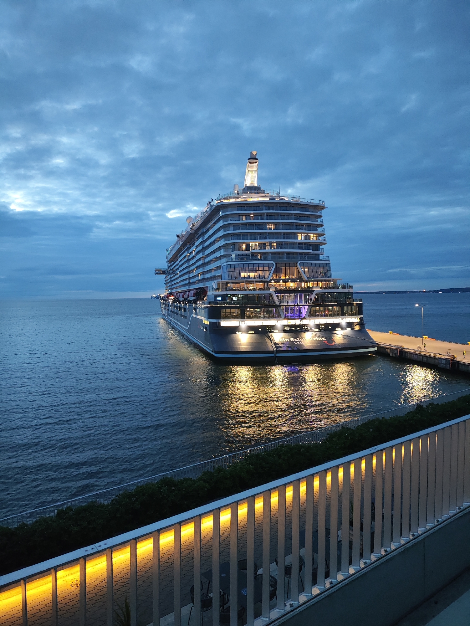 A large cruise ship, identified by the text "EIN SCHIFF RELAX" on its stern, is docked at a pier in Tallinn, Estonia. The ship is illuminated, with lights visible in many of its windows. The sky is overcast with clouds, and the sea is calm. In the foreground, a white railing with integrated yellow lighting runs along a walkway or balcony overlooking the water and the ship. The overall scene is of a port at dusk or early evening.