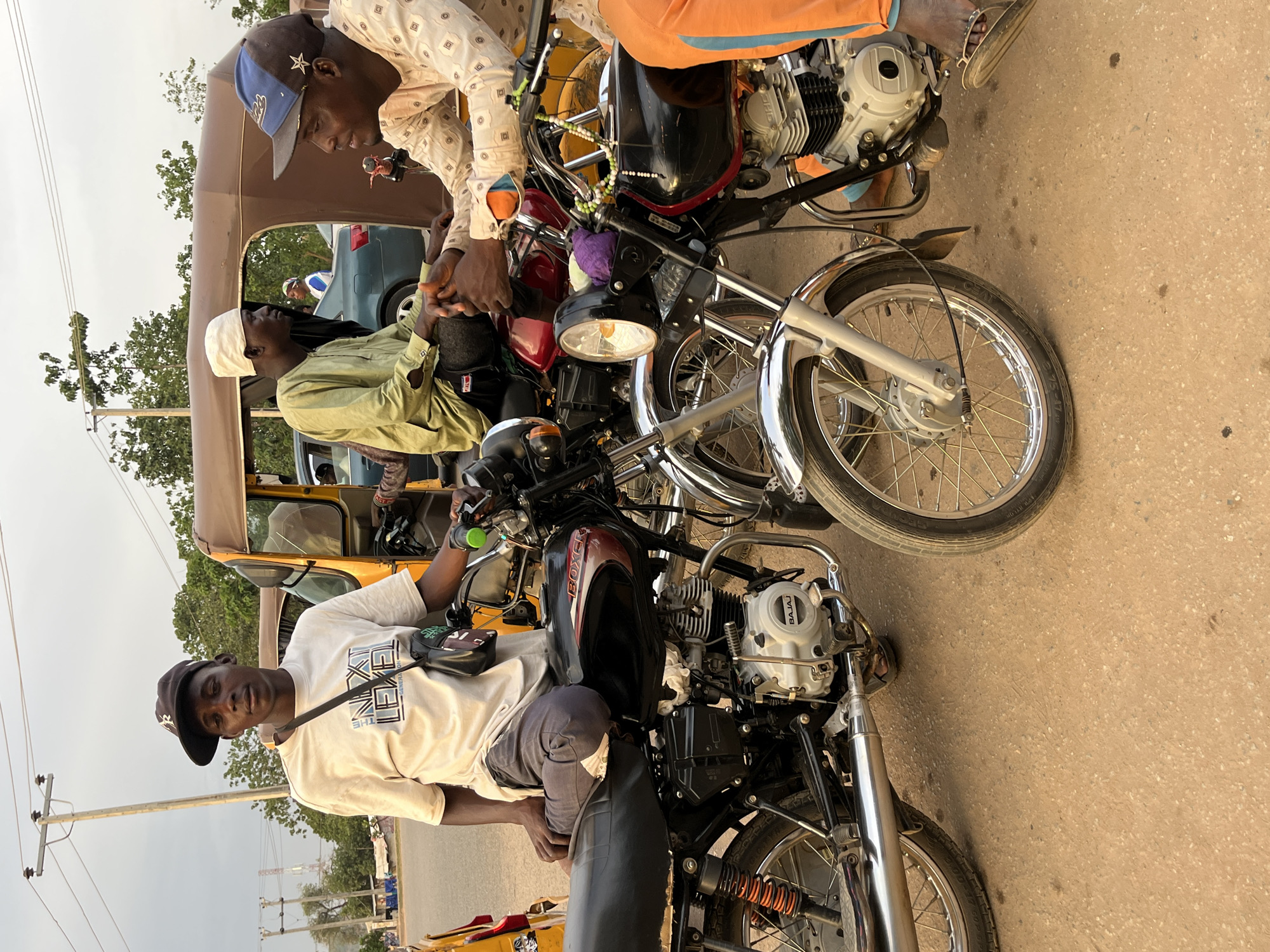 This image captures a daytime street scene in Abuja, Nigeria, featuring several men and vehicles. The sky is overcast or hazy, suggesting either an early morning, late afternoon, or cloudy day.
In the foreground, a young man wearing a white t-shirt with "THE NEXT LEVEL" printed on it and a black baseball cap sits astride a black and red motorcycle, looking directly at the camera with a relaxed expression. He has a black shoulder bag across his chest. This motorcycle has "BOXER" on its fuel tank and "BAJAJ" visible on its engine casing.
To the right of him, another motorcycle is positioned, partially obscured. Two men are engaged in conversation around this motorcycle and a nearby yellow/brown tricycle (commonly known as a "keke napep"). One man, wearing a patterned long-sleeved shirt and a blue baseball cap, stands leaning over the handlebars of the motorcycle. He appears to be talking to a man seated inside the tricycle, who is wearing a light green shirt and a white cap. Beads are strung around the handlebars of the motorcycle next to the standing man.
In the background, behind the tricycle, a blue or grey car is visible on the road. Power poles with multiple utility lines and sparse trees line the street, indicating an urban or semi-urban environment. The ground appears to be a light brown, possibly dirt or unpaved road. The overall atmosphere suggests a casual gathering or a brief stop along a thoroughfare.