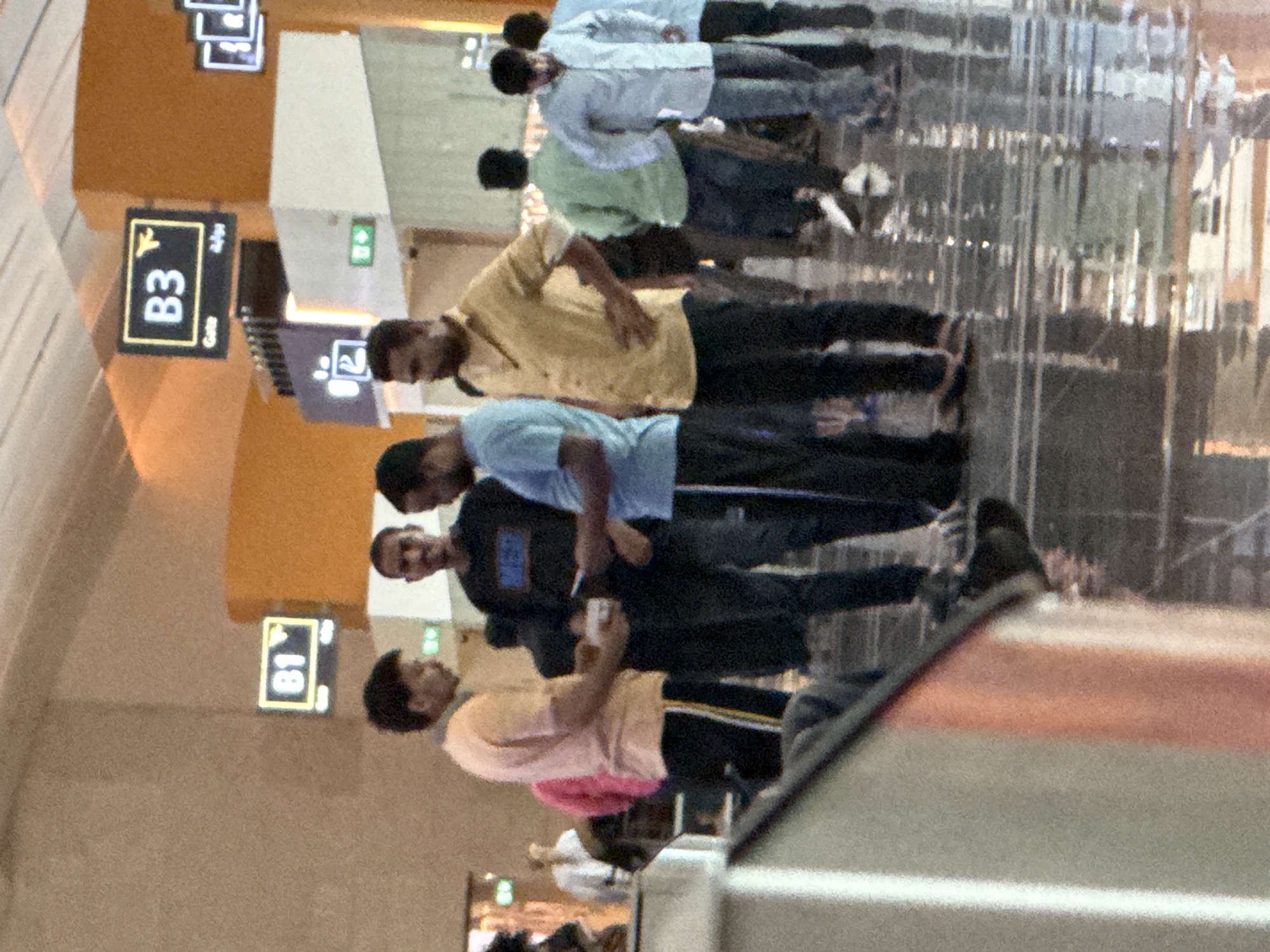 The image depicts a group of men standing in what appears to be an airport terminal in Seeb Province, Oman. Several overhead signs are visible, including one displaying "B3" and an airplane icon, and another labeled "B1". The floor is highly reflective, suggesting a polished surface, and the lighting indicates an indoor setting. The men are casually dressed and appear to be engaged in conversation or waiting. One man in the foreground is wearing a light pink shirt and dark pants. Another man next to him is in a dark shirt and dark pants. To his right, a man in a yellow shirt and dark pants is standing with his hands on his hips. Further back, other individuals are visible, some in green and light blue shirts, also appearing to be waiting or moving through the terminal. The overall atmosphere is that of a busy public transit hub.
