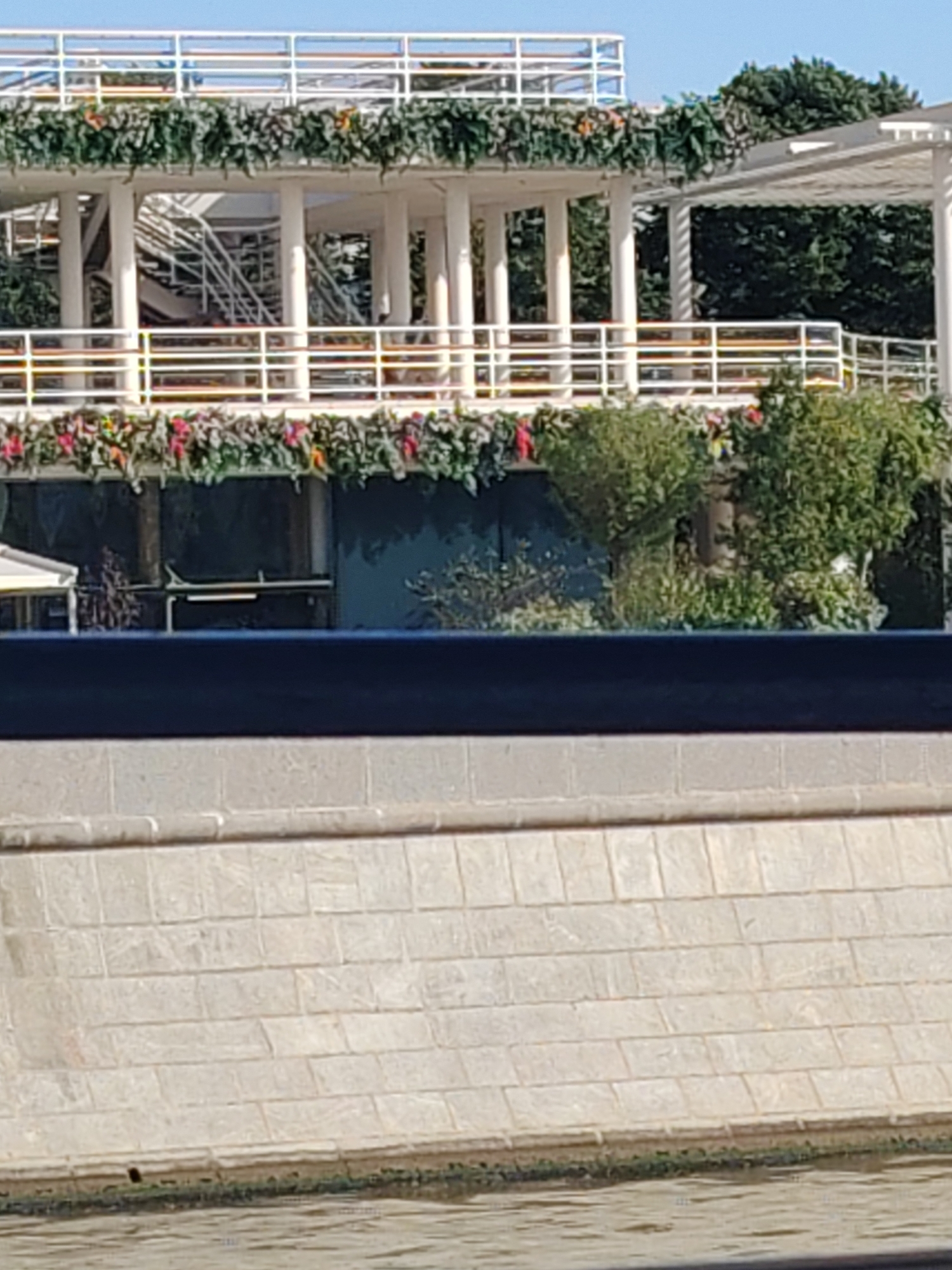 A white, multi-level structure, possibly a building or pier, is decorated with garlands of green foliage and red flowers. The structure has white railings and columns, and stairs are visible within its levels. The background is filled with lush green trees and a clear blue sky. In the foreground, a light-colored stone wall lines a body of water, with some dark, possibly algae, growth at the waterline. The lighting suggests it is daytime.