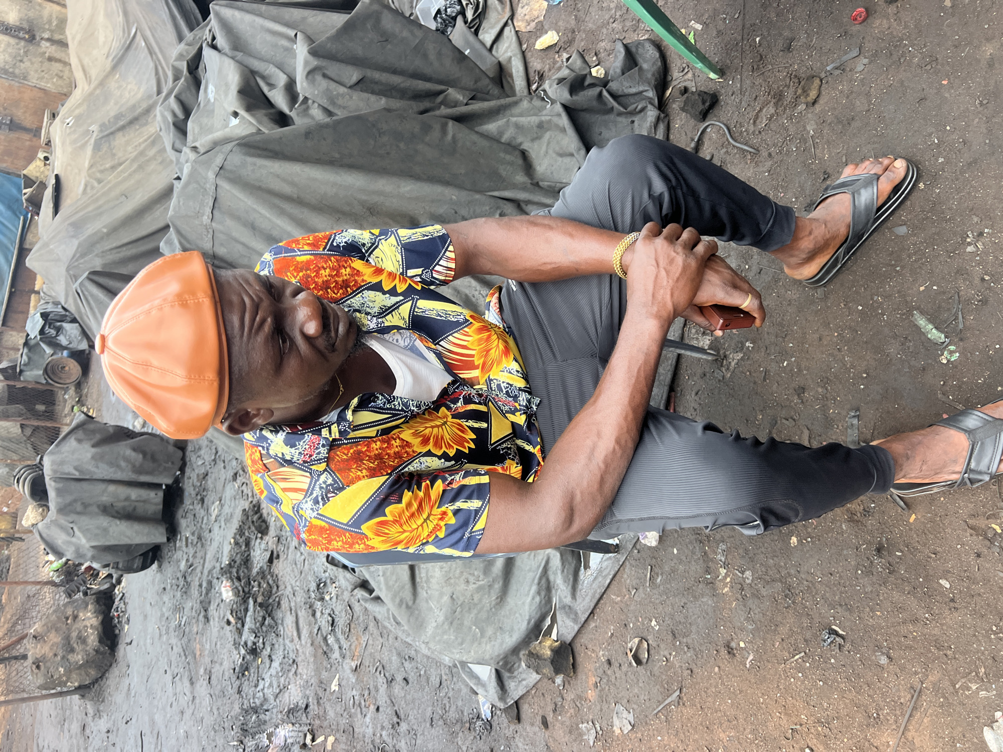 A man is sitting on the ground outdoors, with his legs extended and bent at the knees. He is wearing a vibrant short-sleeved shirt with a pattern of yellow and orange flowers and a white undershirt. On his head, he wears an orange flat cap, and on his feet, black flip-flops. A gold bracelet is visible on his left wrist. His hands are clasped, and he appears to be holding a small, dark rectangular object. He is looking down, with a pensive expression.
The setting is a gritty, outdoor area. The ground is dirt and debris, with scattered bits of metal and trash. In the background, large, dark gray tarps are draped over what appear to be piles of objects or vehicles. The scene suggests a working environment, possibly a market or a repair yard, in Obosi, Nigeria. The lighting indicates it is daytime, and the weather appears to be overcast or dusty. There is no visible text in the image.