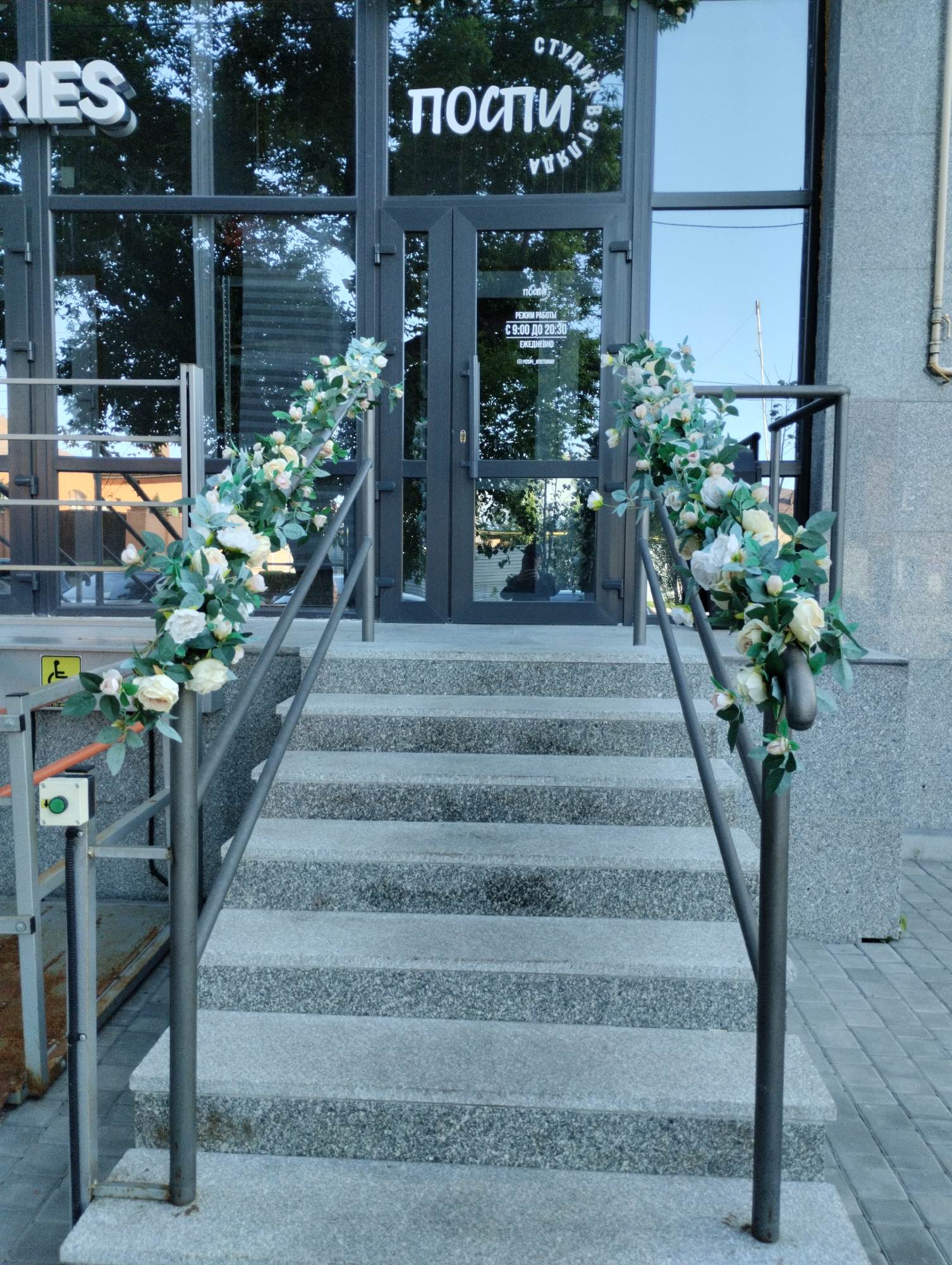 The image shows a building entrance with a concrete staircase and railings decorated with artificial flowers. Above the entrance, text is visible on the glass doors, which appears to be in Cyrillic script. One part of the text reads "ПОСТИ" and another part, possibly the name of the establishment, is "СТУДИЯ КРАСОТЫ" or similar. Below this, smaller text indicates operating hours: "РЕЖИМ РАБОТЫ C 9:00 ДО 20:30 ЕЖЕДНЕВНО" (Opening hours from 9:00 to 20:30 daily). A social media handle "@kesha_kostanay" is also visible. The architectural style suggests a modern building. The presence of Cyrillic script and the social media handle mentioning "kostanay" confirms the location as Kostanay, Kazakhstan. The scene is captured during daylight hours, with natural light illuminating the exterior of the building. There are no people visible in the immediate vicinity of the entrance.