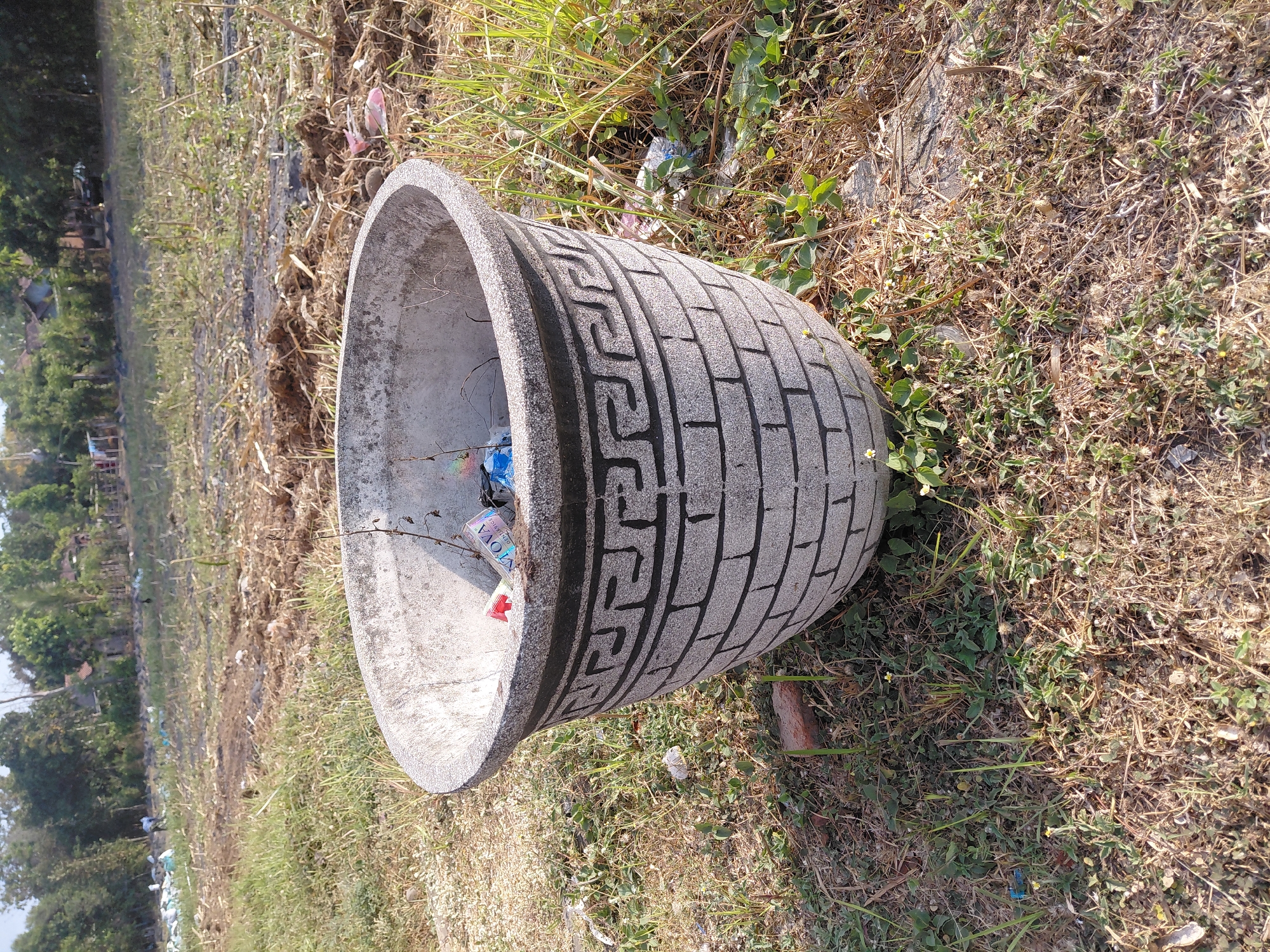 A weathered, grey concrete planter sits in an outdoor, grassy area. The planter is decorated with a black, Greek key pattern and a brick pattern. Inside the planter, there is a small amount of debris, including what appears to be cigarette packs and plastic bottles. The ground surrounding the planter is covered in dry grass, some green vegetation, and scattered bits of trash. In the background, there is a field with patches of bare earth and more debris. Further in the background, trees and a few buildings are visible, suggesting a semi-rural or edge-of-town setting in Alas Tengah, Indonesia. The sky is clear and the lighting suggests it is daytime.