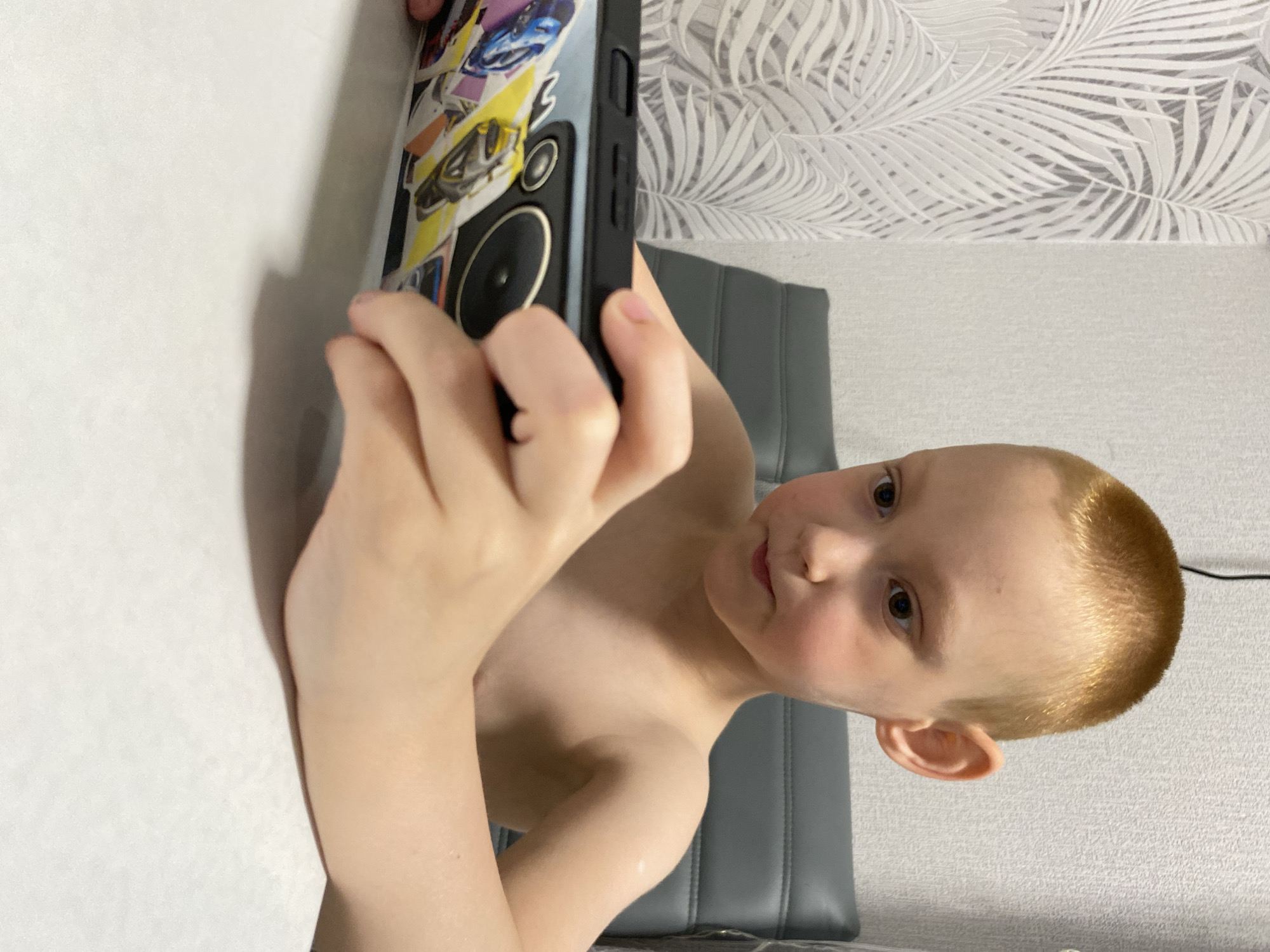 A young boy, appearing to be between 4-7 years old, is depicted in an indoor setting. He has short, light brown or blonde hair and light-colored eyes, and is shirtless. His expression is neutral to slightly curious as he looks directly at the camera; his cheeks are faintly flushed.
He holds a smartphone or tablet horizontally with both hands. The device has a black protective case featuring multiple colorful stickers or a vibrant patterned design. These patterns include abstract shapes and stylized images, resembling racing cars in purple and yellow. A distinct circular element, likely a camera lens or design feature, is visible on the phone's rear.
The boy is seated, with a dark grey, padded surface, possibly a chair or headboard, visible behind him. A plain white table or surface occupies the foreground, extending from the left and partially obscuring his lower body and arms. The background consists of wallpaper adorned with a repeating pattern of light grey or white palm fronds against a slightly darker grey base. A thin black cable is visible against the wallpaper in the upper right.
The scene appears to be an interior space, likely a home in Noviy, Russia. The lighting is soft and ambient, providing no specific cues about the time of day. No discernible text is visible within the image.