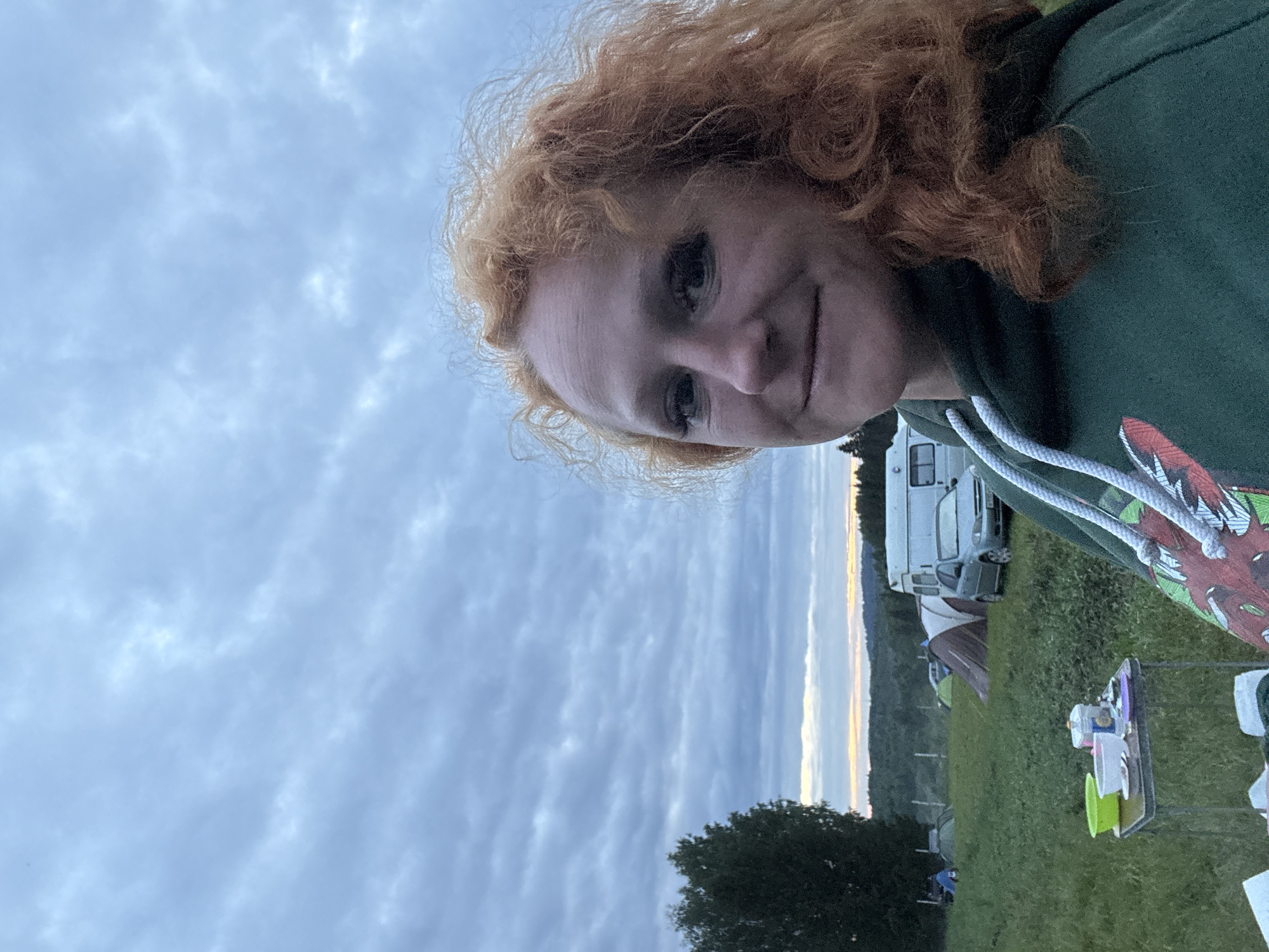 The image captures a candid outdoor scene in Большая Кивара, Russia, during twilight or an overcast day. A woman, with curly reddish-orange hair, is positioned in the right foreground, looking towards the camera with a slight smile. She is wearing a dark green hooded sweatshirt with white drawstrings and a partially visible red and white graphic design on the chest. To her left, a small, foldable camping table holds a bright green bowl, a white bowl, a clear bottle, and various dishes. The midground reveals a grassy field with multiple tents, including a brown and tan tent, a white campervan, and a light-colored sedan. In the background, forested hills are visible under an overcast sky with textured grey clouds. A narrow band of faint orange light is present on the distant horizon, and a large, dark green tree stands on the far left, with two indistinct figures visible near its base.