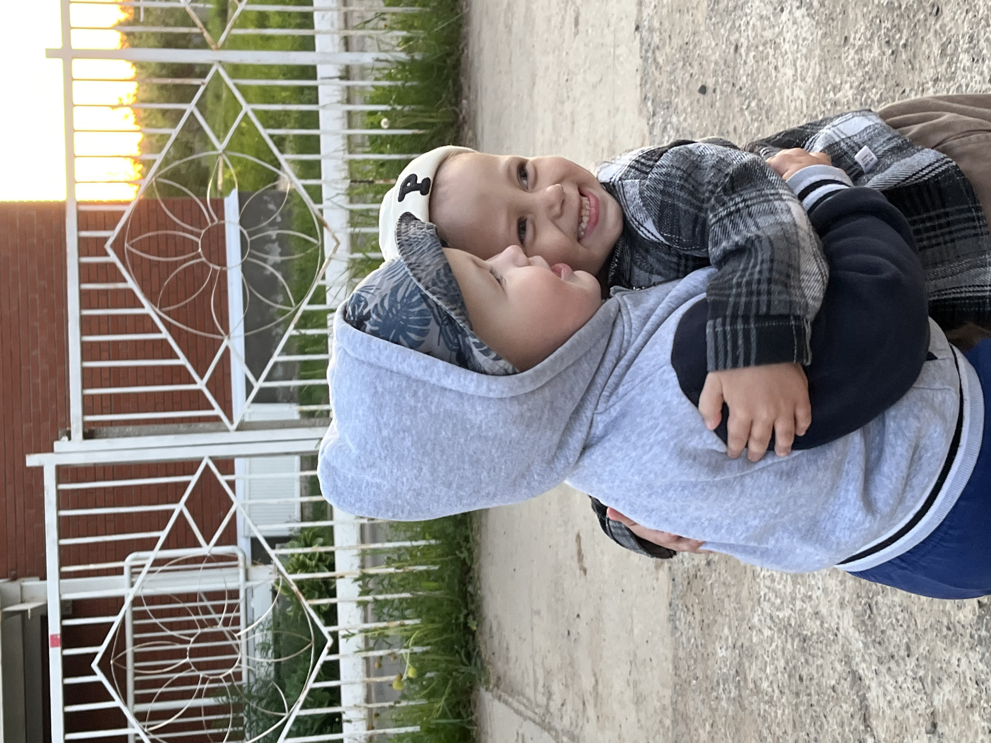 Two young children are embracing outdoors in Фоки, Russia. One child wears a gray patterned hooded sweatshirt over a navy blue garment with white stripes, while the other child wears a plaid shirt and a white cap. They are on a concrete surface. Behind them is a white decorative metal fence. Sunset lighting is visible in the upper left corner.