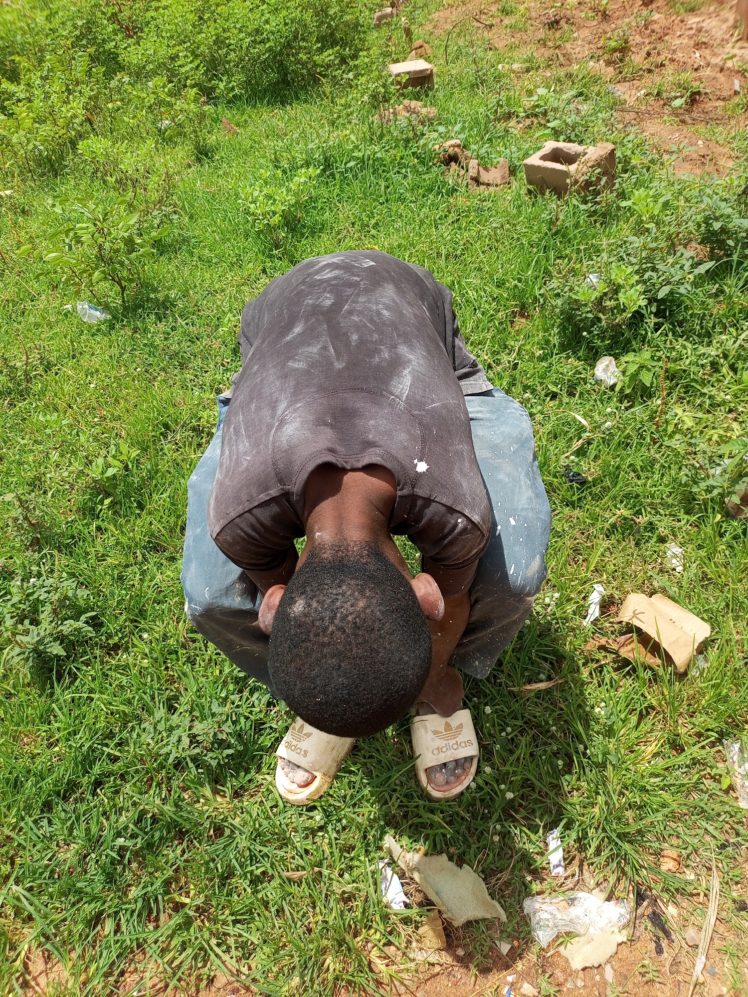 A person is crouching on the ground in an outdoor area with a lot of grass and some dirt. The person is wearing a dark shirt with white splatters and blue pants. They are bent over, with their head down. There are some scattered objects on the ground, including a piece of cardboard and some debris. In the background, there are some bricks and more green vegetation. The scene appears to be in an undeveloped or construction area.