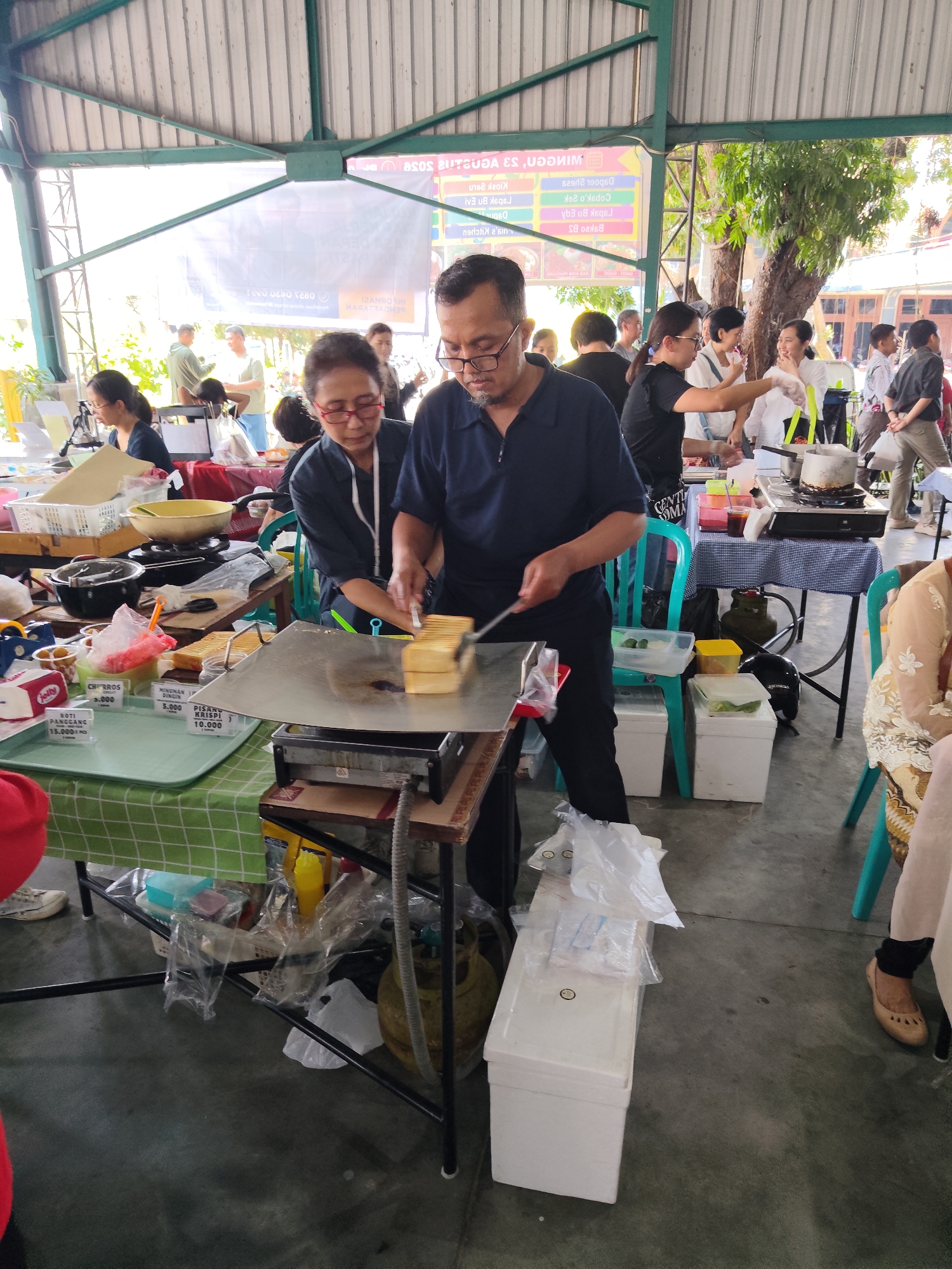 In Malang, Indonesia, at a market or outdoor food stall event, a middle-aged couple is preparing food. The man, wearing glasses and a navy blue polo shirt, is focused on grilling several slices of bread on a flat-top griddle. To his left, a woman with glasses and a dark blouse is also attending to the food preparation. They are working at a wooden stall with a green and white checkered tablecloth. On the table, various food items and containers are visible, including plastic bags of ingredients, a yellow bowl, and what appear to be pre-packaged snacks. In the background, other individuals are seen milling around the market, with a large banner in Indonesian hanging above. The environment appears to be outdoors, possibly under a covered structure, with trees and other market stalls in the distance.