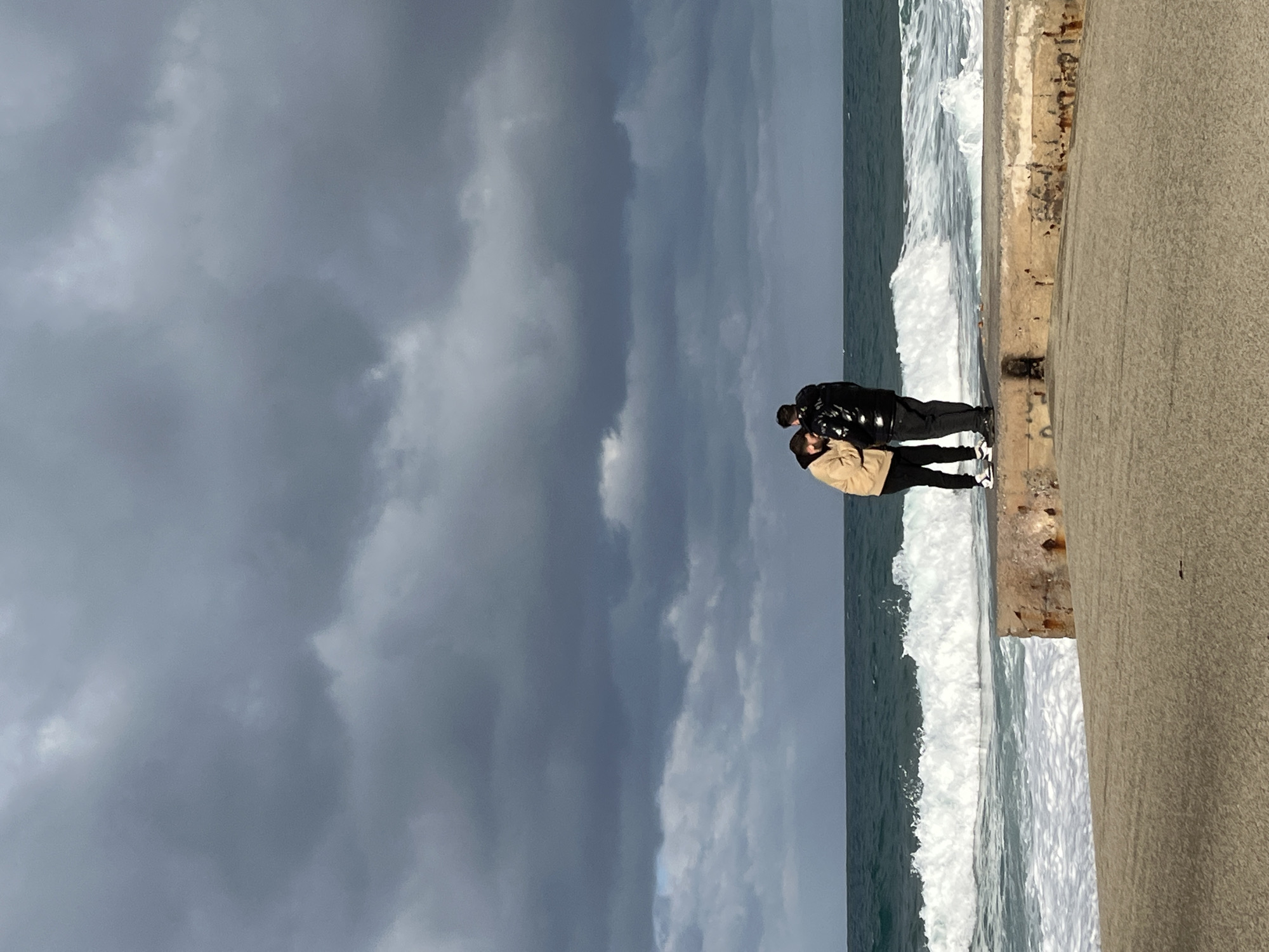 Two people stand on a concrete pier next to the ocean in Alanya, Turkey. Dark storm clouds fill the sky. One person is wearing a tan jacket; the other, a black jacket. White waves crash against the pier.