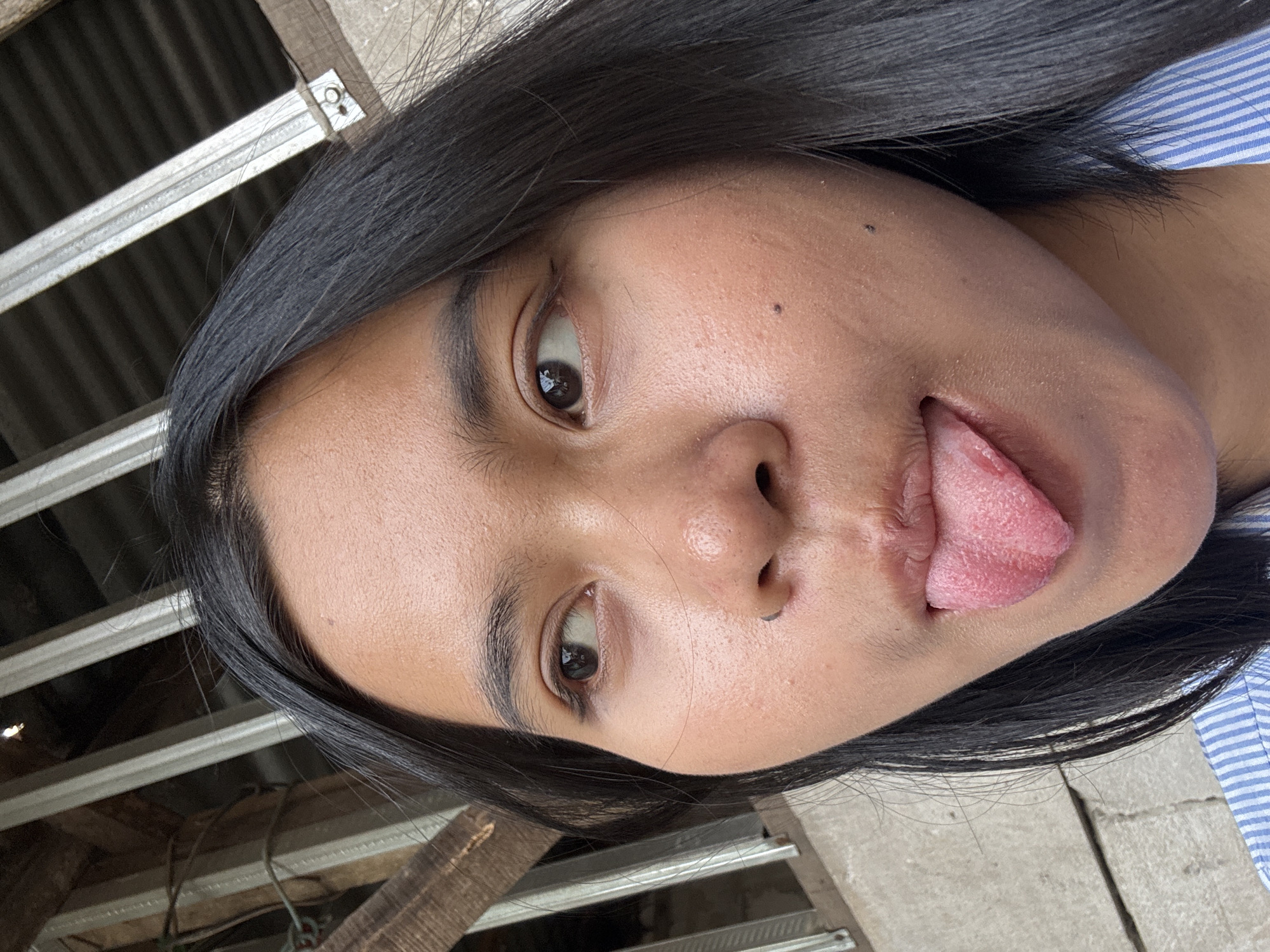 A close-up, eye-level shot shows a young Southeast Asian woman sticking out her pink, textured tongue. Her dark hair frames her face as she looks to her left. She has a light complexion with a few beauty marks visible on her cheeks and nose. The background features weathered wooden planks and corrugated metal roofing, suggesting an outdoor setting that could be part of an informal residential structure in East Jakarta, Indonesia. The woman is wearing a blue and white striped collared shirt.