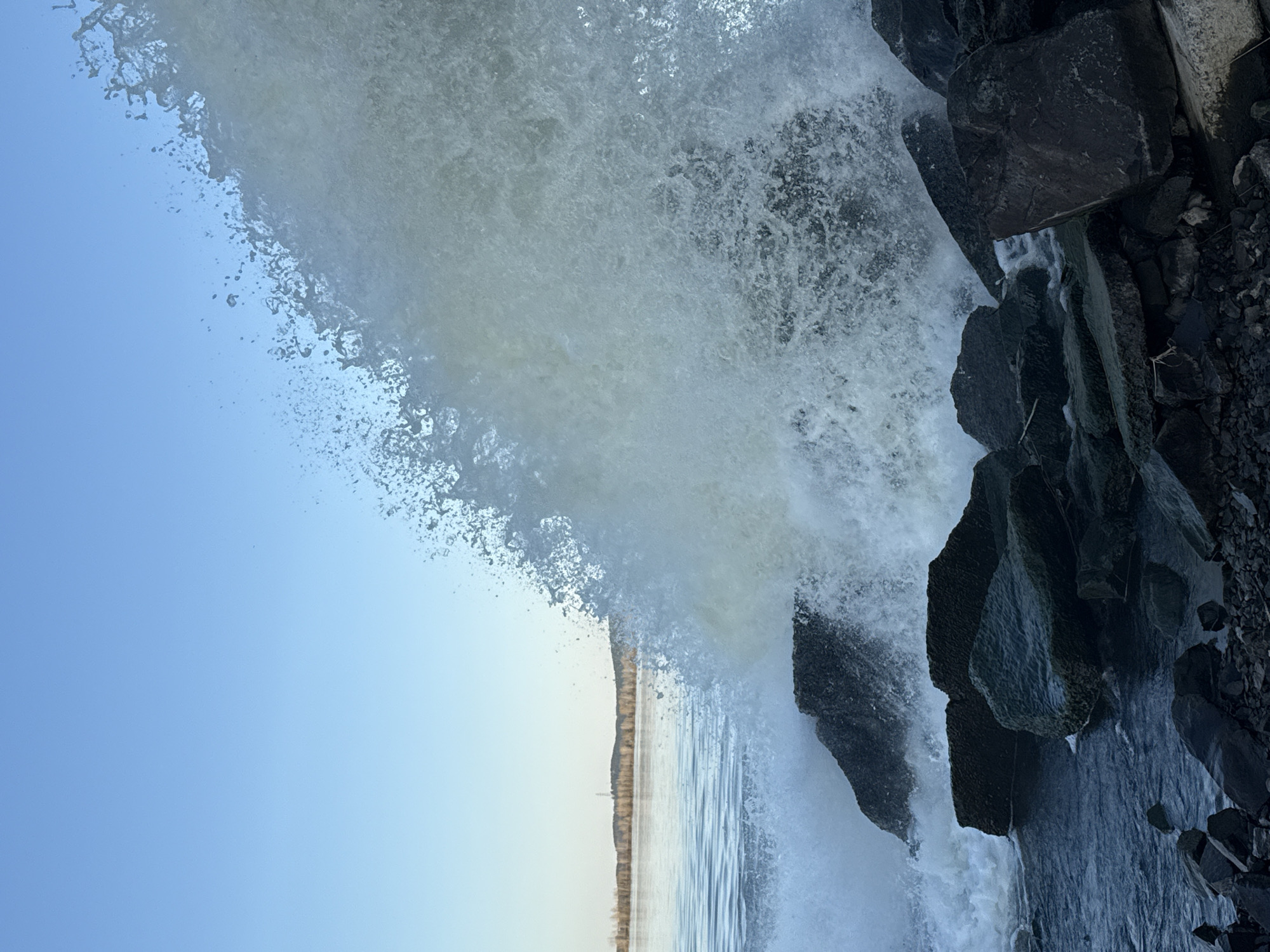 A large wave crashes against a rocky shore. The water spray is mostly white and opaque, partially obscuring the view of the sky and a distant shoreline. The sky is blue. The rocks are dark and appear wet. The location in Chernushka, Russia, cannot be verified based on the image alone.