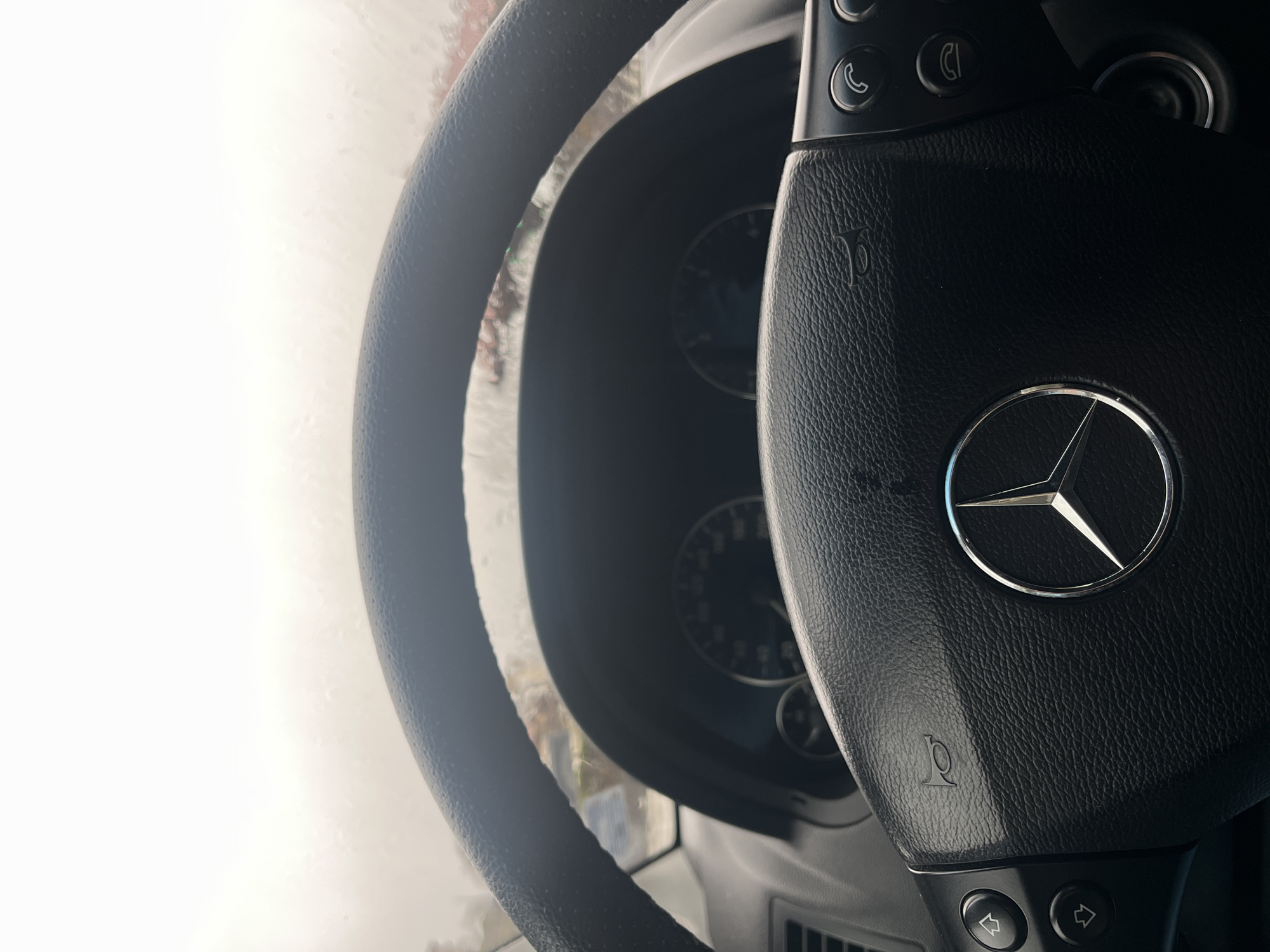 The image shows the interior of a Mercedes-Benz vehicle. The steering wheel is prominently displayed with the Mercedes-Benz logo visible in the center. Controls are on the wheel. The dashboard is slightly blurred in the background, with partial view of a speedometer. The car is wet, presumably from rain. The exact location in Srednogortsi, Bulgaria, cannot be determined based solely on the provided image.