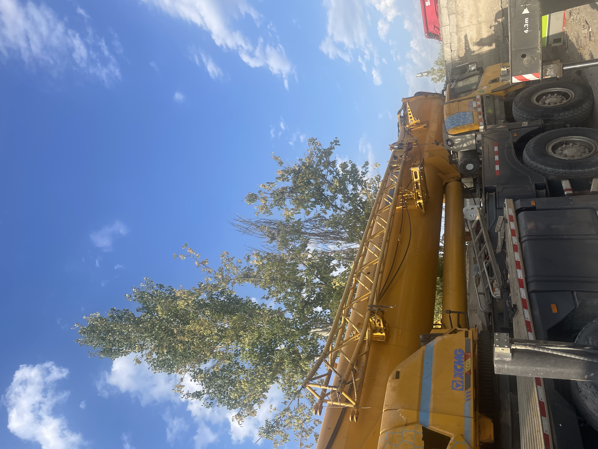 A large yellow mobile crane, identified by the "XCMG" logo, is parked outdoors. The crane's telescopic boom is extended and rests above the vehicle's cabin. Behind the crane, several trees with sparse green and yellowing leaves are visible against a bright blue sky with scattered white clouds. The ground appears to be unpaved, with some debris present. To the right of the crane, a concrete wall is visible, and a red object, possibly another piece of equipment, is partially visible in the background. A sign on the crane's outrigger indicates "4.3m". The scene suggests a construction or industrial setting. The image does not contain any people or animals.