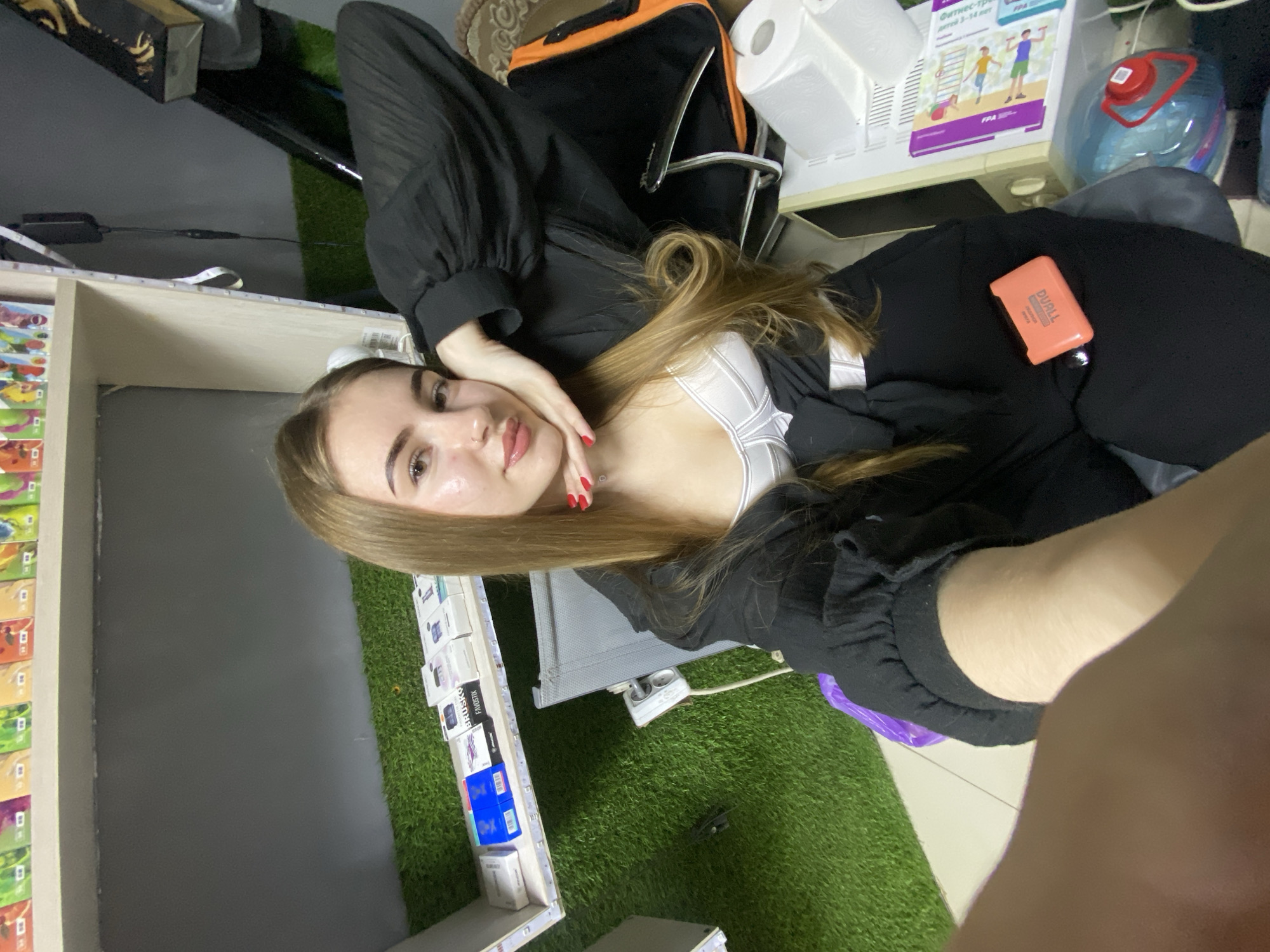 The image depicts a young woman with long, blonde hair and fair skin, wearing a white bralette and a black open cardigan. She is sitting in what appears to be an indoor setting, possibly a store or a room with shelves displaying various products. The shelves are stocked with numerous small boxes, some of which are labeled. In the background, there is artificial grass covering a section of the wall or floor, suggesting a decorative element. To the right of the woman, a desk or table is visible, with items such as a roll of toilet paper, a water cooler bottle, and some papers with colorful illustrations. An orange object, possibly an electronic device, is placed on her black clothing. The overall lighting suggests an indoor environment, with some artificial lighting present. The specific location of Дьяково, Russia, is not explicitly visible or identifiable from the image content alone.
