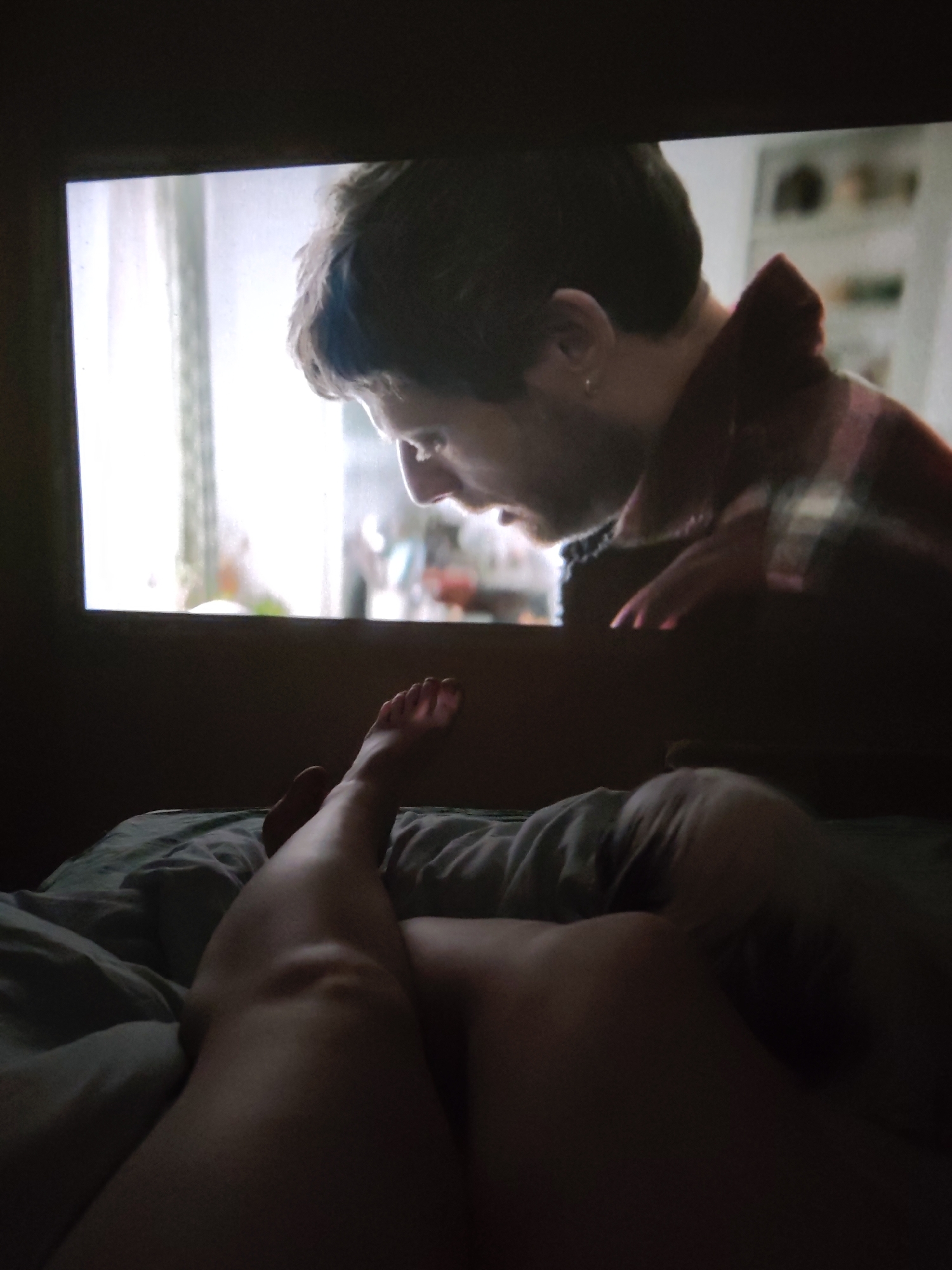 A person is lying in bed, covered by a teal-colored comforter. Their legs are visible, with one foot extended. A screen displaying a person in a reddish-brown flannel is in the background. The location is not evident.