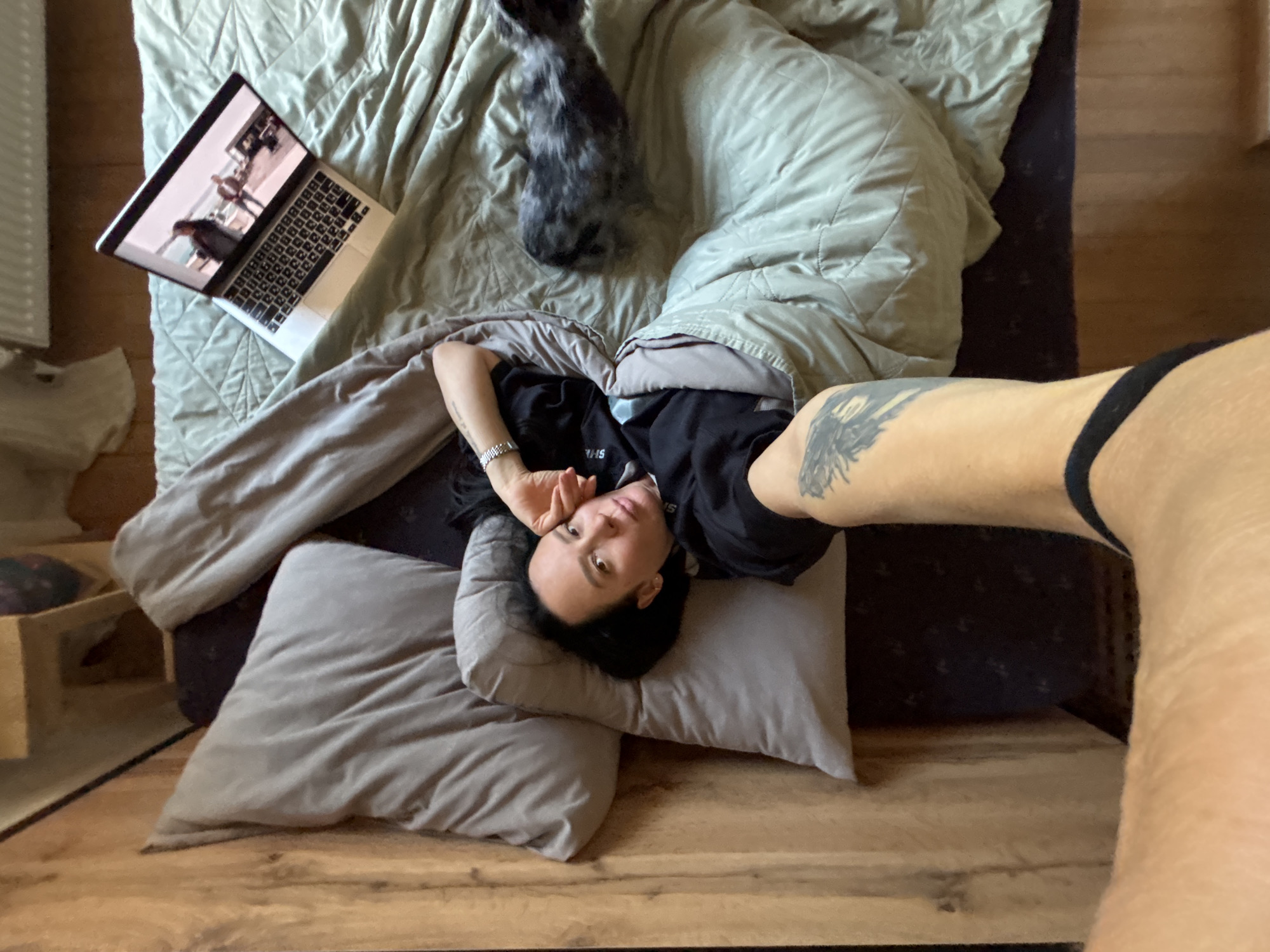A woman lies on a bed with a laptop and a gray cat. She is lying on her side with a pillow under her head and a blanket partially covering her. She has a tattoo on her left arm. The laptop is on the bed beside her and appears to be playing a video. The cat is lying near her head, also on the bed. A portion of the woman's leg is visible in the lower right corner of the image. The setting appears to be indoors, possibly a bedroom. The location is Chaikovsky, Russia.