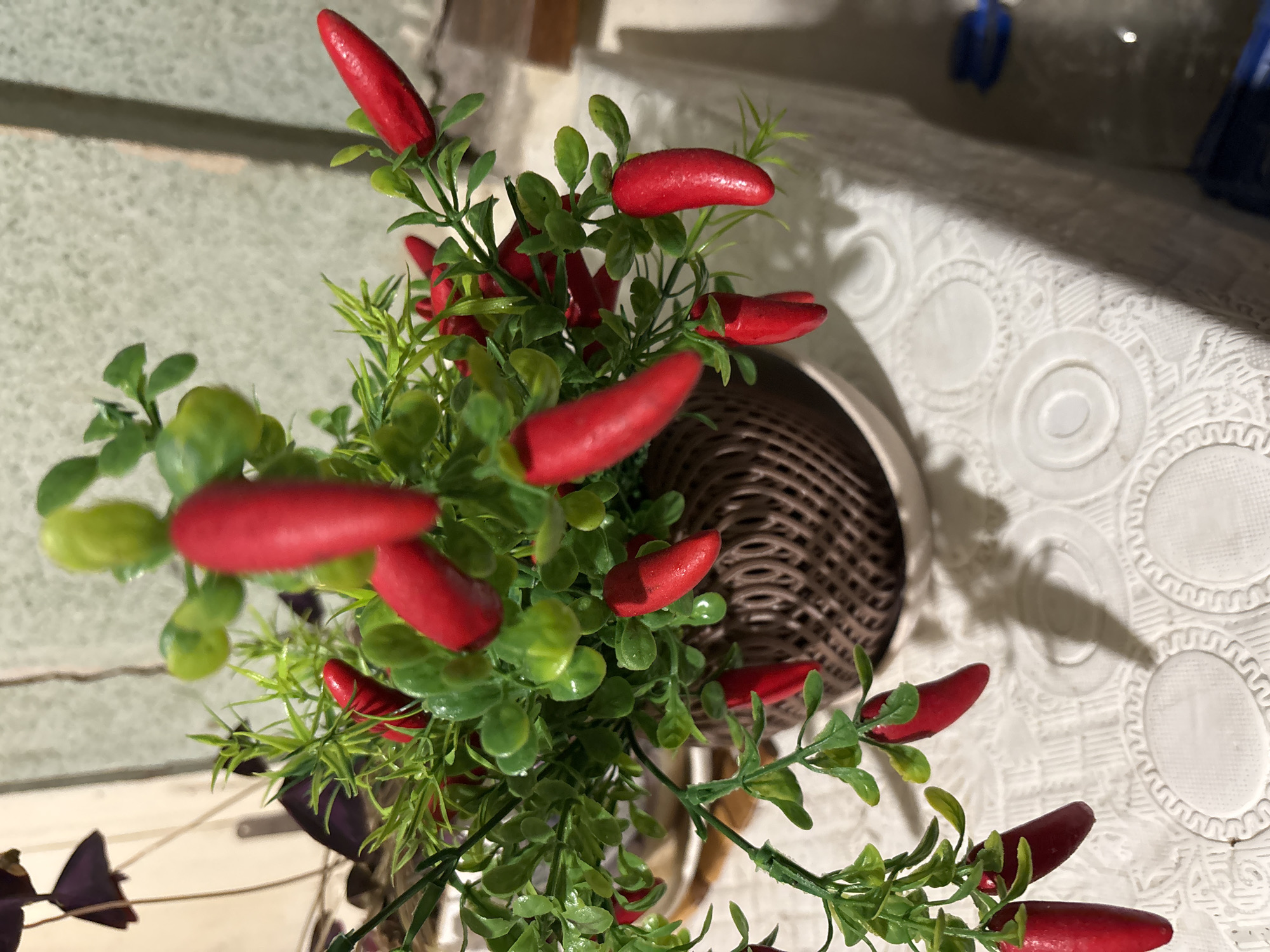 This overhead shot showcases a vibrant artificial plant featuring numerous bright red, chili-pepper-shaped fruits and glossy green leaves. The plant is situated in a dark brown, woven pot or basket, prominently visible on the right side of the frame. It serves as a decorative centerpiece in what appears to be an indoor setting.
The plant rests on a white tablecloth, which displays an intricate embossed pattern of concentric circles and wavy designs. Shadows are cast on the tablecloth, suggesting a daytime setting with a light source from above or to the side. In the blurred background on the tablecloth, indistinct objects, possibly blue in color, can be faintly seen in the upper right. To the left, a blurry light-colored surface and a dark, wing-shaped decorative element are visible. The plant itself is in sharp focus, while the background elements exhibit a shallow depth of field. No specific activity is depicted, and no text is visible in the image. The scene likely takes place on a table inside a home or establishment in Vagharshapat, Armenia.