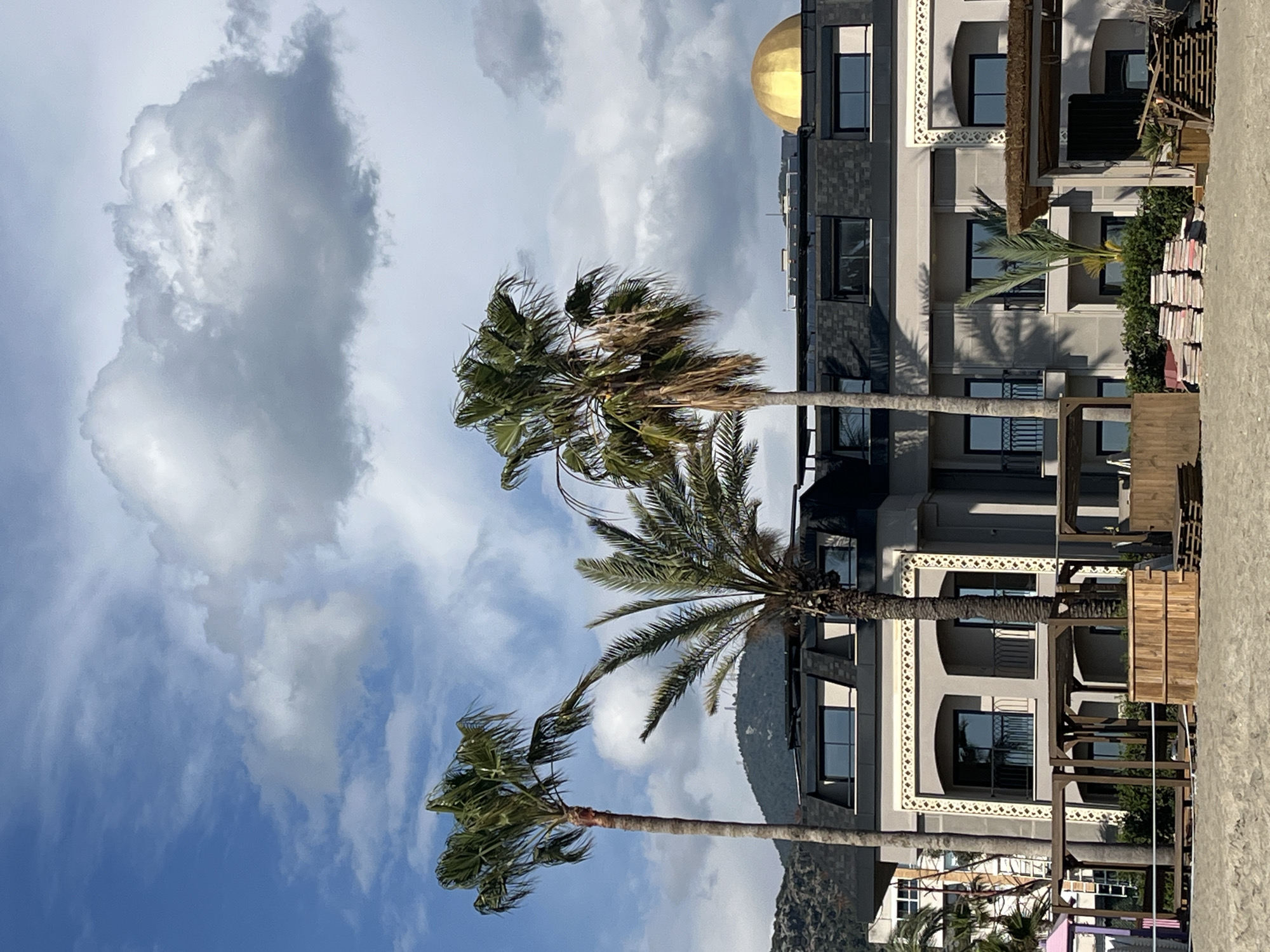 The image displays a beachfront scene in Alanya, Turkey, centered on a multi-story building and several palm trees. The foreground consists of light-colored sand. On the sand are two partially constructed or dismantled wooden pergolas, stacks of reddish-pink square cushions, and stacks of wooden pallets. A small wooden planter containing a young palm tree is also present.
The main structure is a grey and beige building, likely a hotel or resort, featuring multiple dark-framed windows, some with black metal balcony railings. Distinctive white and gold geometric trim adorns sections of the building's facade. The roof is dark grey and mansard-style, equipped with dormer windows and visible solar panels. A prominent golden dome is situated on the right side of the roofline. Green bushes are visible at the base of the building.
Three palm trees of differing heights stand in front of and beside the building, displaying green fronds. The background features forested mountains with structures visible on their peaks. The sky is partly cloudy, with areas of blue showing through scattered white and grey cumulus clouds. No individuals are present in the scene.
