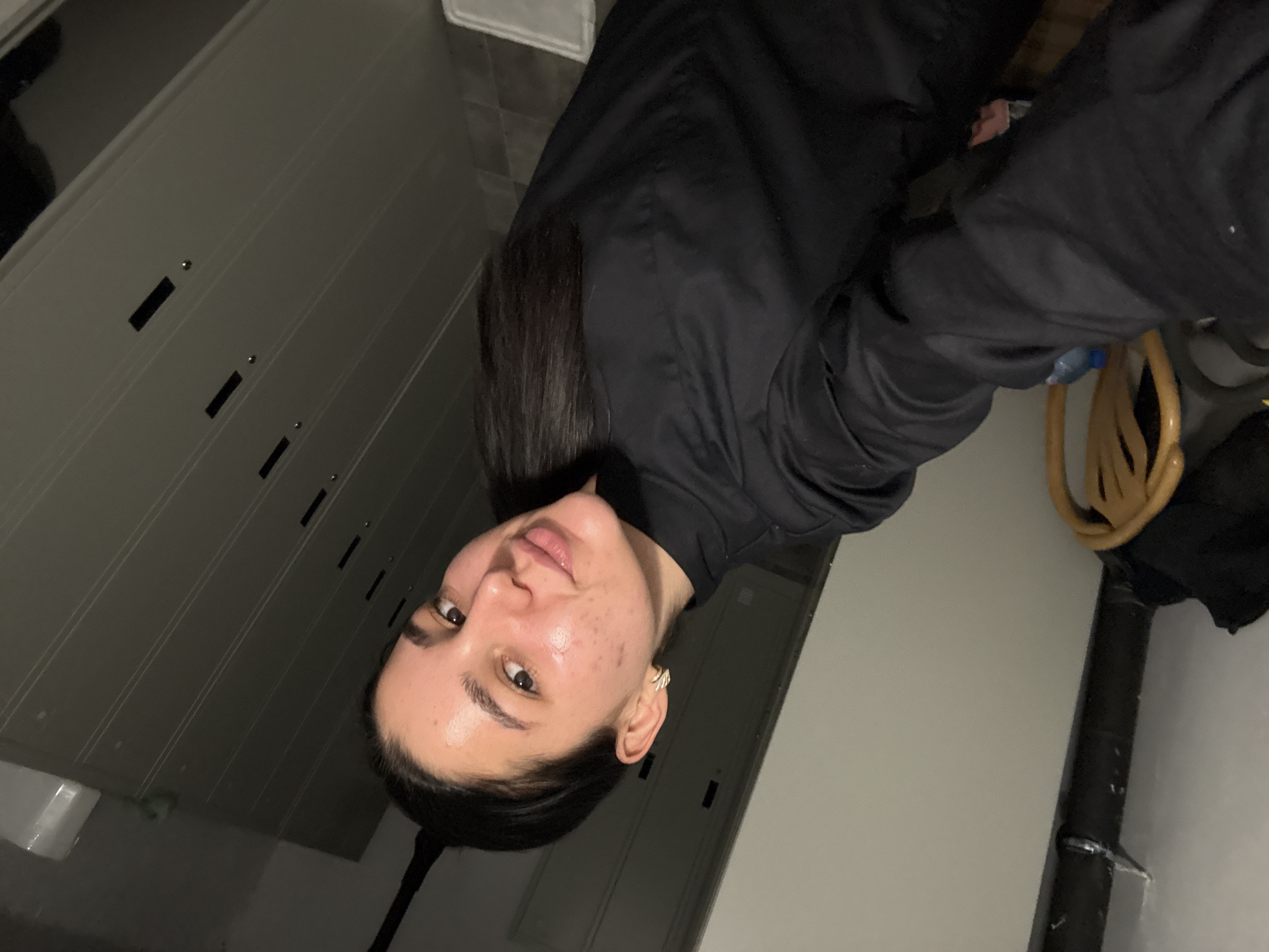 A woman with dark hair, wearing a black jacket, is posing for a selfie. The background appears to be an interior space with what seems to be a wall with built-in storage or lockers. A chair with a woven back and a black object are also visible. It is not possible to determine a location in Veliko Tarnovo, Bulgaria, based on the visual information in the image.