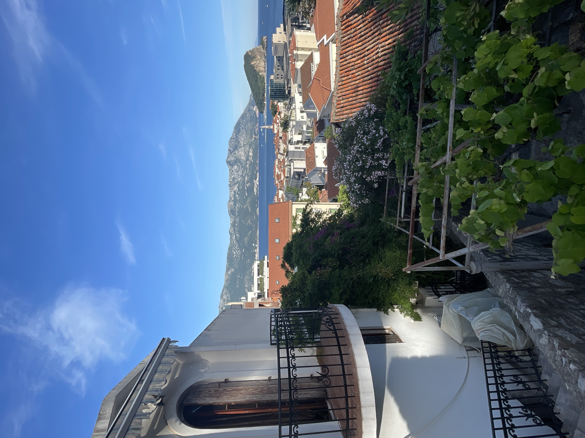 The image shows a view overlooking the city of Budva, Montenegro, captured from an elevated position. The scene includes:
**Environment:** A bright, sunny day with a blue sky dotted with light clouds. The Adriatic Sea is visible in the background, with mountains and coastal structures lining the distant horizon.
**Buildings and Structures:** The foreground contains parts of buildings, particularly a white structure with a curved arch and a black wrought-iron railing. Terracotta rooftops of other buildings spread into the middle ground, interspersed with greenery.
**Vegetation:** There is abundant greenery, including vines and flowering bushes, that frame parts of the scene.
**Objects:** A metal trellis-like structure is visible among the greenery. A portion of what appears to be a covered or wrapped object sits near the white building.
**Location:** Likely taken from an elevated residential area in Budva, providing a panoramic view of the city, coastline, and sea.