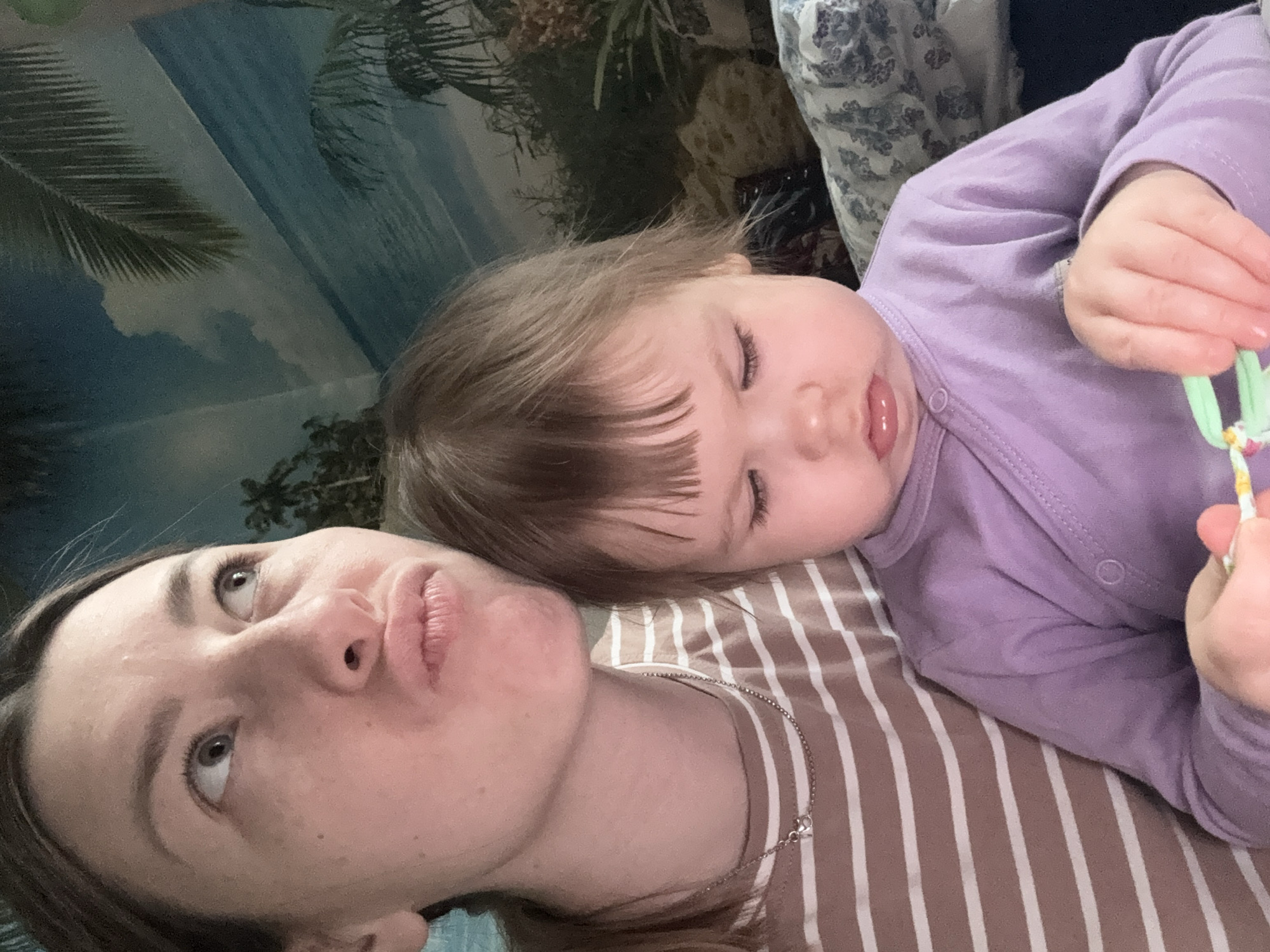 A woman and a baby are lying down. The woman has light skin, brown hair, and is wearing a brown and white striped shirt. She is looking up and to the left. The baby, lying on the woman's chest, has light skin, brown hair, and is wearing a purple onesie. The baby's eyes are mostly closed, and their tongue is slightly protruding. The background includes a wall with a beach scene and palm tree decorations. The location of the image is unknown; the claim to be in Krapivka, Russia is not supported by any details in the image.