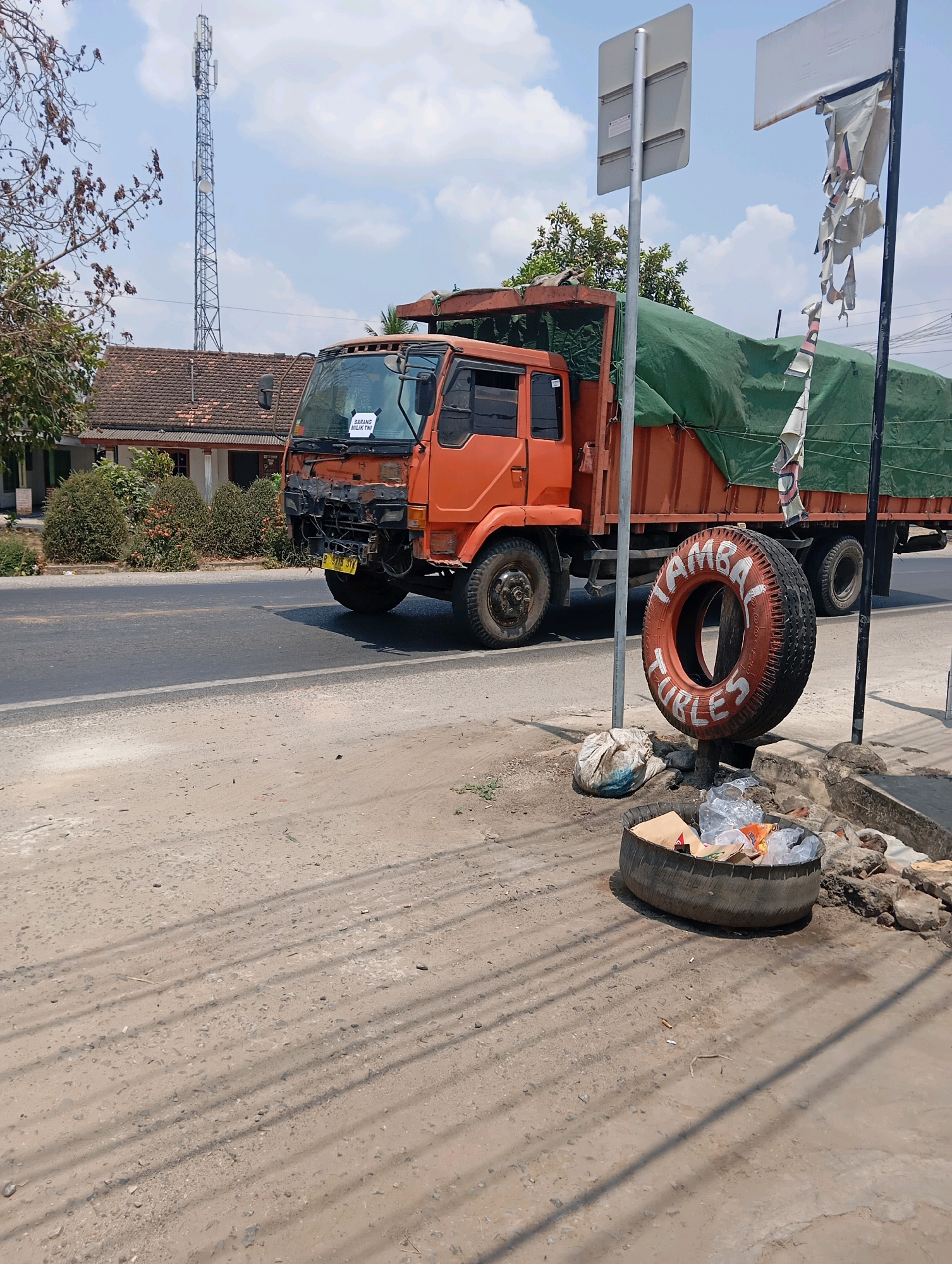 An orange dump truck is parked on the side of a road. The truck's front bumper is damaged and missing. A green tarp covers the truck bed. A spare tire, painted white with red lettering that reads "TAMBAL BAN (TIRE REPAIR)", is mounted vertically on a post near the truck. Below the tire is a tire repurposed as a trash bin, filled with plastic bags and other debris. In the background, a telecommunications tower stands tall against a blue sky with scattered clouds. A residential building with a tiled roof is visible behind the truck. The ground is made of dirt and gravel, with tire tracks imprinted on it. The scene appears to be in a rural or semi-rural area, possibly near Leman Benawi, Indonesia, given the context of the question.