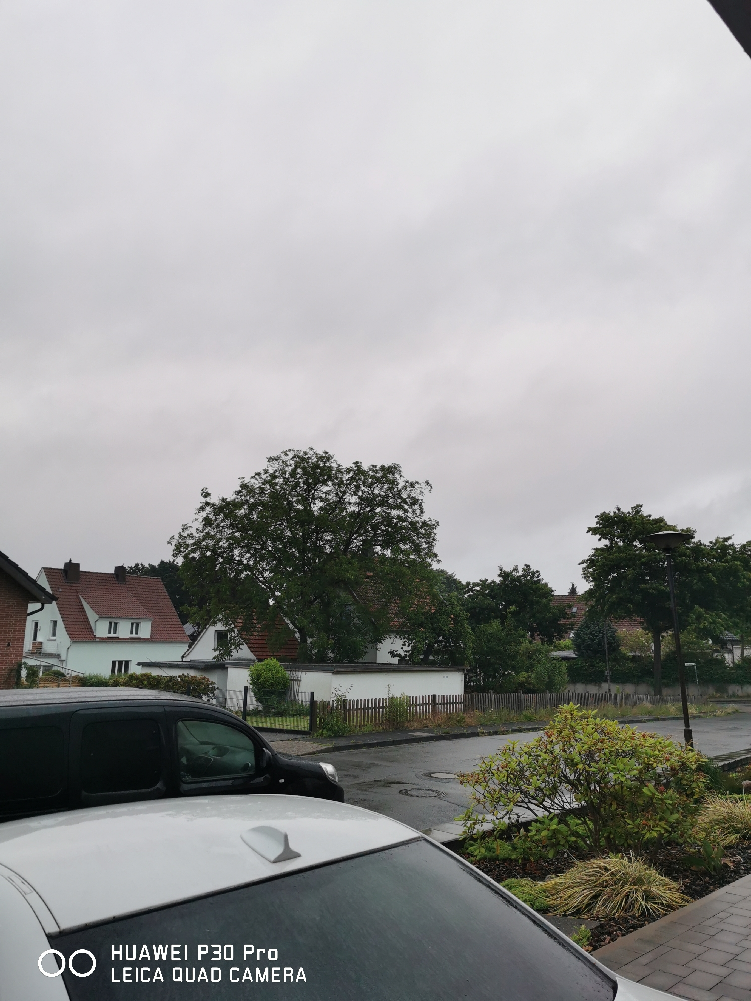 The image shows a cloudy day in a residential area. In the foreground, the roof of a white car is visible, with a shark fin antenna and water droplets on its surface, indicating recent rain. Behind the white car, a black van is parked, its windows tinted. The van is parked on a wet asphalt road, with manhole covers visible. Beyond the road, a wooden fence separates the properties from a garden with a small tree and decorative plants. Further back, there are several houses with red-tiled roofs and light-colored walls, surrounded by mature trees. A street lamp is also visible on the right side of the image. The overall impression is a suburban street scene on an overcast day.