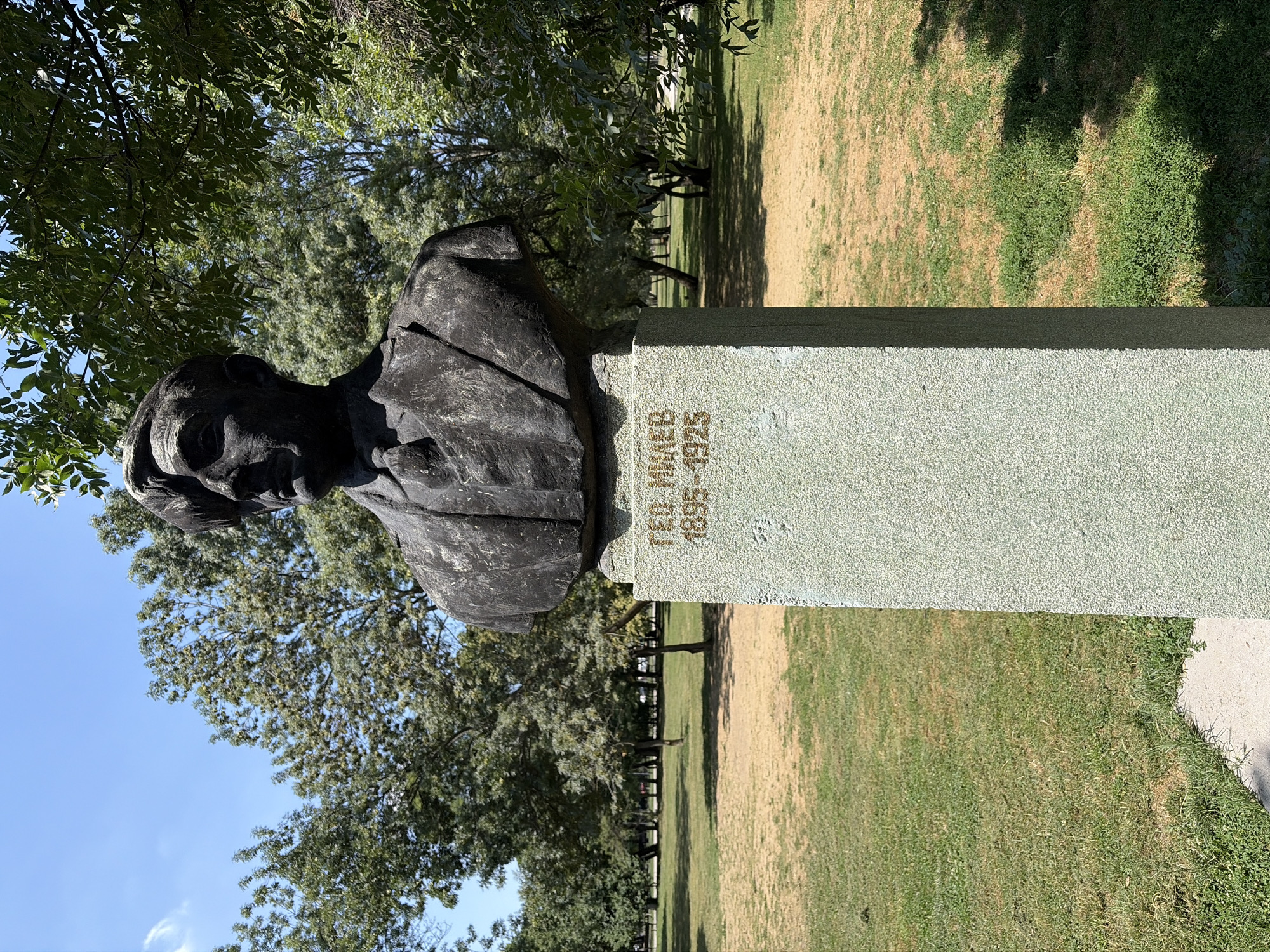 A bust of a man, likely Geo Milev, is situated on a stone pedestal in a park. The bust is dark in color and depicts the man from the chest up, wearing a suit and tie. The pedestal is light gray and has inscriptions in gold letters. The inscription reads "ГЕО МИЛЕВ 1895-1925". The background consists of a grassy area with scattered trees and a clear blue sky with some clouds. The scene is outdoors and appears to be a sunny day. Based on the inscription, this monument is likely located in Sofia, Bulgaria, honoring the Bulgarian poet and writer Geo Milev.