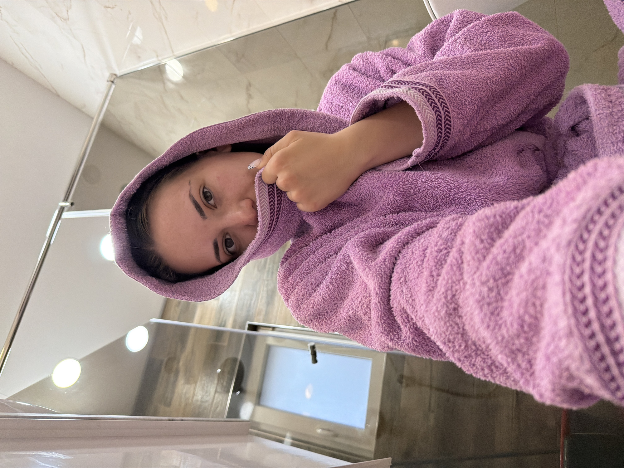 A young woman wearing a purple terrycloth bathrobe with a hood is looking at the camera. Her hand is raised near her face. The setting appears to be a bathroom, with white marble walls, a window, and a stainless-steel shower frame visible. The location may be a bathroom in Buhovo, Bulgaria.