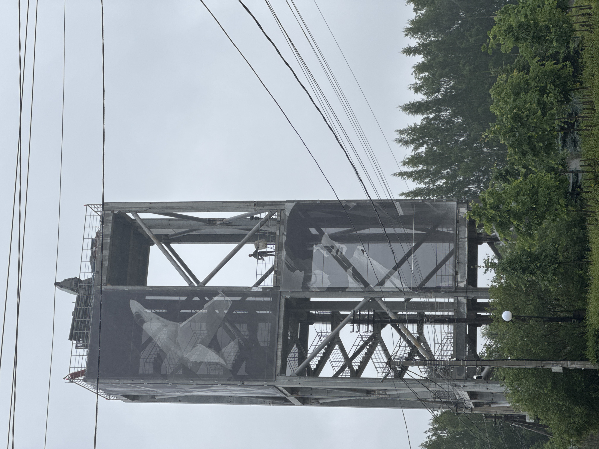 A tall, industrial concrete tower is pictured against a cloudy sky. The tower has a lattice-like structure and is partially covered by a large tarp featuring a graphic of a fighter jet. Two figures, likely workers, are visible on a platform near the top of the tower, one of whom appears to be holding a tool. Staircases are visible ascending the interior of the tower. In the foreground, lush green trees and foliage are present, and a lamppost with a spherical light fixture stands among the greenery. Electrical wires crisscross the upper portion of the image. The scene suggests an urban or industrial setting with surrounding nature.