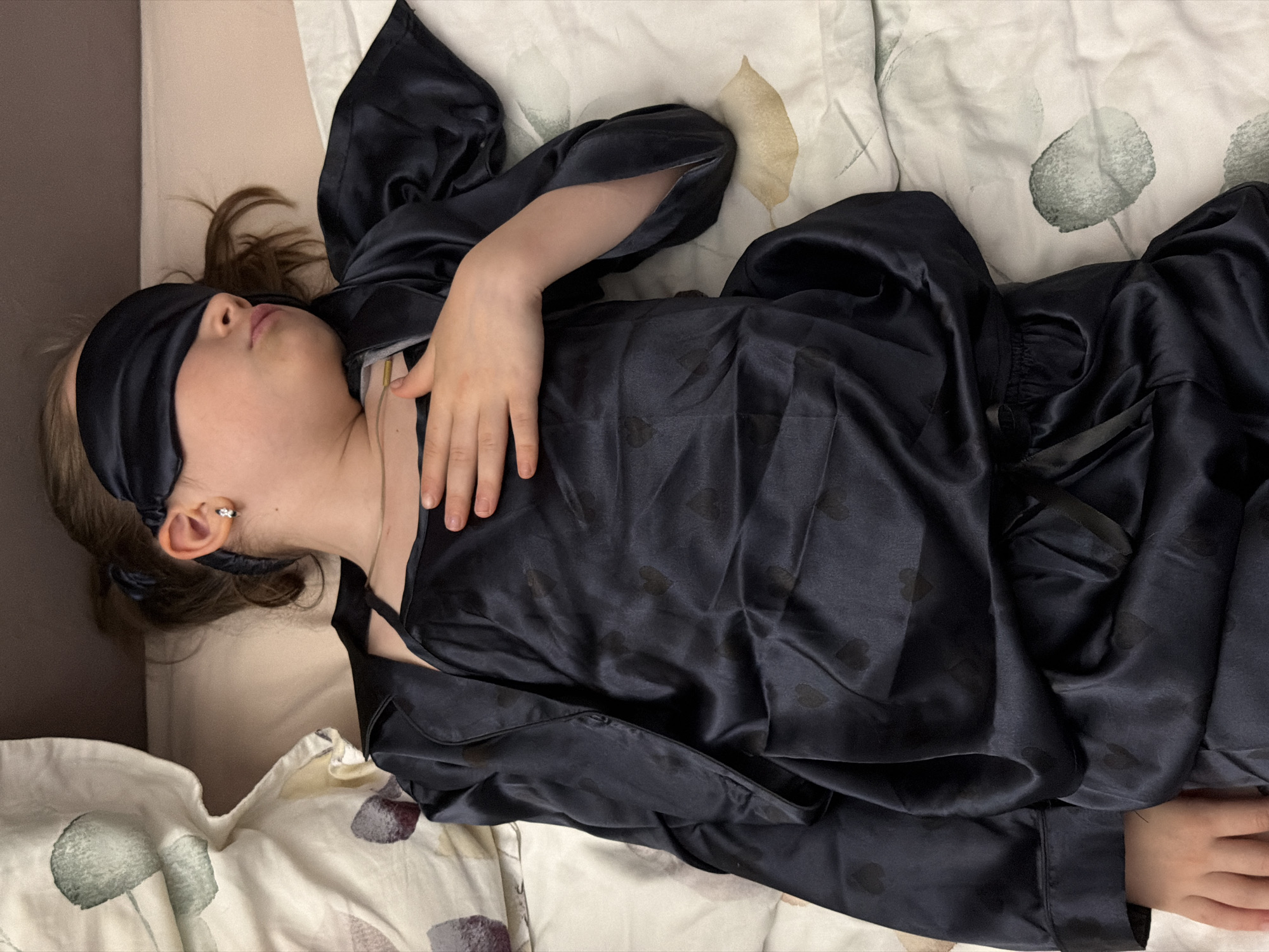 A child lies in bed wearing a dark blue silk pajama set and a matching sleep mask. The child is lying on their back on a bed with patterned sheets. The background includes a plain headboard. The location is the city of Perm, Russia.