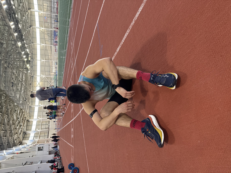 An individual is seated on a red indoor running track within a large athletics facility in Kazan, Russia. The male subject, appearing to be in his twenties, has short dark hair and is wearing a light blue and white gradient sleeveless tank top, black shorts, red socks, and dark blue running shoes with white and yellow soles. A black sports watch is on his left wrist, and a black heart rate monitor is strapped to his right bicep. He is positioned with his elbows resting on his knees, hands loosely clasped, and head lowered, gazing at the track directly in front of him, indicating a state of rest or exhaustion.
In the midground, several other individuals are visible further down the track. A male figure in a grey t-shirt and blue jeans stands with his back to the viewer, observing a small group of athletes in athletic attire who are jogging or walking. A series of red and white hurdles are positioned near these individuals. To the far left, two blue and red circular objects, possibly track and field equipment, are visible on the track's surface.
The environment is a spacious indoor stadium characterized by a high, arched ceiling with exposed metal trusses, ventilation ducts, and numerous overhead lighting fixtures. Large windows or skylights are visible along the upper perimeter. Beyond the track, a green artificial turf field extends across the arena, with additional individuals visible in the far distance on the field or in stands. The track itself is marked with white lines delineating multiple lanes.