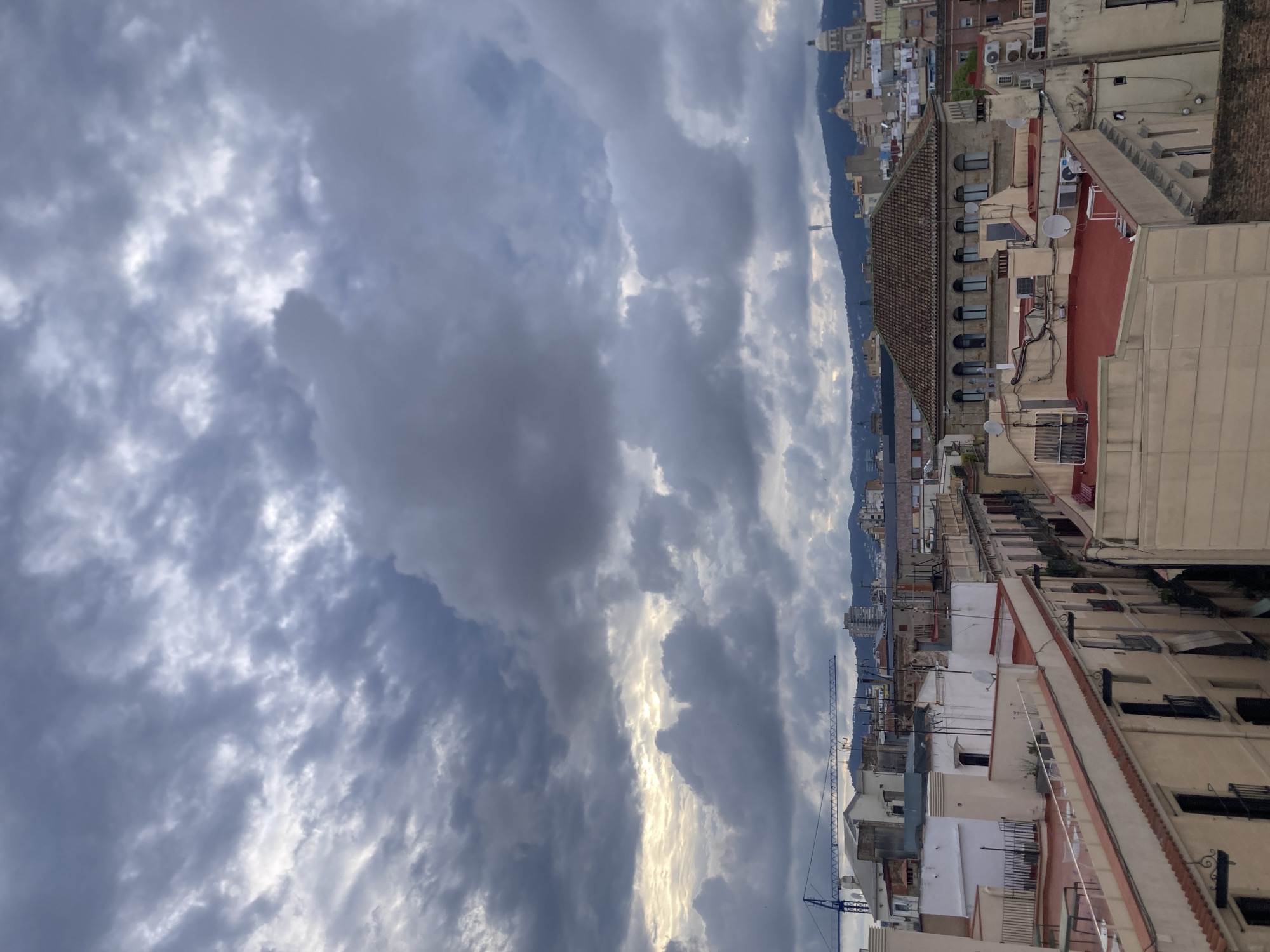 The image depicts a cityscape view, likely in Barcelona, Spain, given the architectural style and presence of a mountain range in the distance. The foreground is dominated by rooftops of buildings, exhibiting a mix of terracotta and light-colored facades. Some buildings have traditional Spanish tile roofs, visible satellite dishes, and balconies. A construction crane is visible in the mid-ground against the backdrop of the city and mountains. The sky occupies a significant portion of the image, featuring dramatic cloud formations with patches of sunlight breaking through. The photograph appears to be taken from an elevated vantage point, looking out over the dense urban landscape towards the distant mountains.