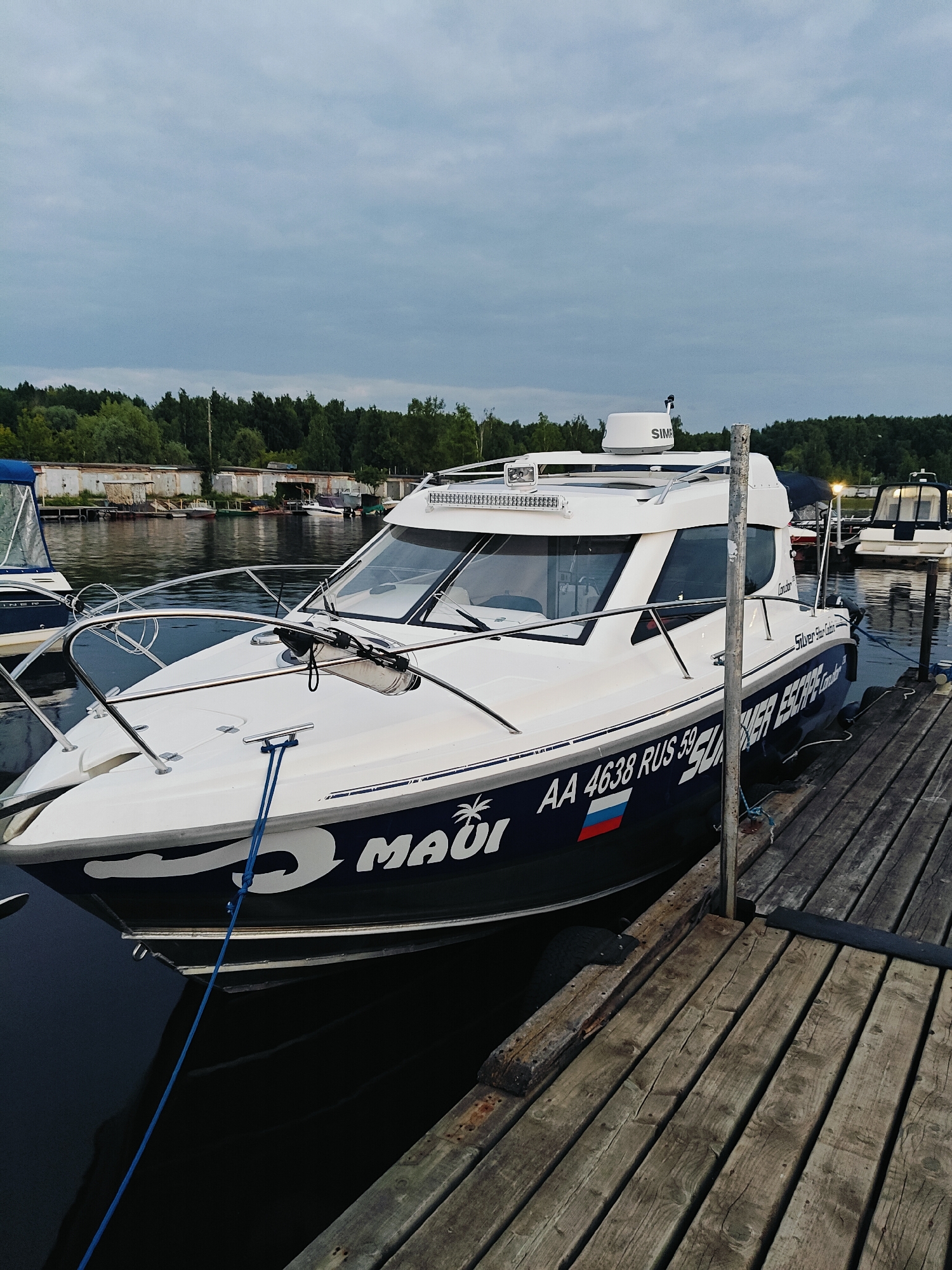 The image captures a white cabin cruiser boat docked at a wooden pier. The boat has "MAUI" and "SILVER ESCAPE" written on its hull, along with the registration "AA 4638 RUS 59". A Russian flag decal is also visible. The boat is secured by a blue rope. In the background, a line of trees and some small buildings are visible along the shore of a body of water, suggesting a marina or waterfront area. The sky is overcast. This scene is likely in the city of Chaikovsky, Russia, given the Russian registration on the boat and the presence of a pier and waterway.
