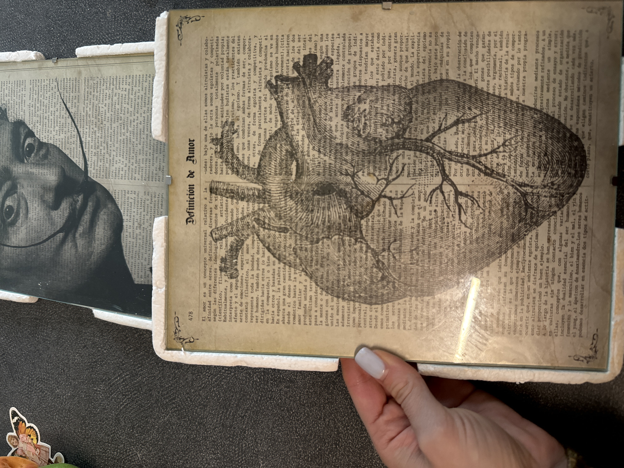 A close-up, overhead shot shows a collage featuring a distressed, sepia-toned heart illustration overlaid with Spanish text and a black and white vintage photograph of a man with a mustache, likely Salvador Dalí. The collage is framed with white textured material, possibly foam or cardboard, and rests on a dark, textured surface, possibly a table or countertop. A small, colorful butterfly sticker is visible in the upper left corner, outside the main framed collage. The lighting appears to be natural, coming from above and to the right, casting a slight glare on the surface of the collage. There is no discernible city or location information present in the image.