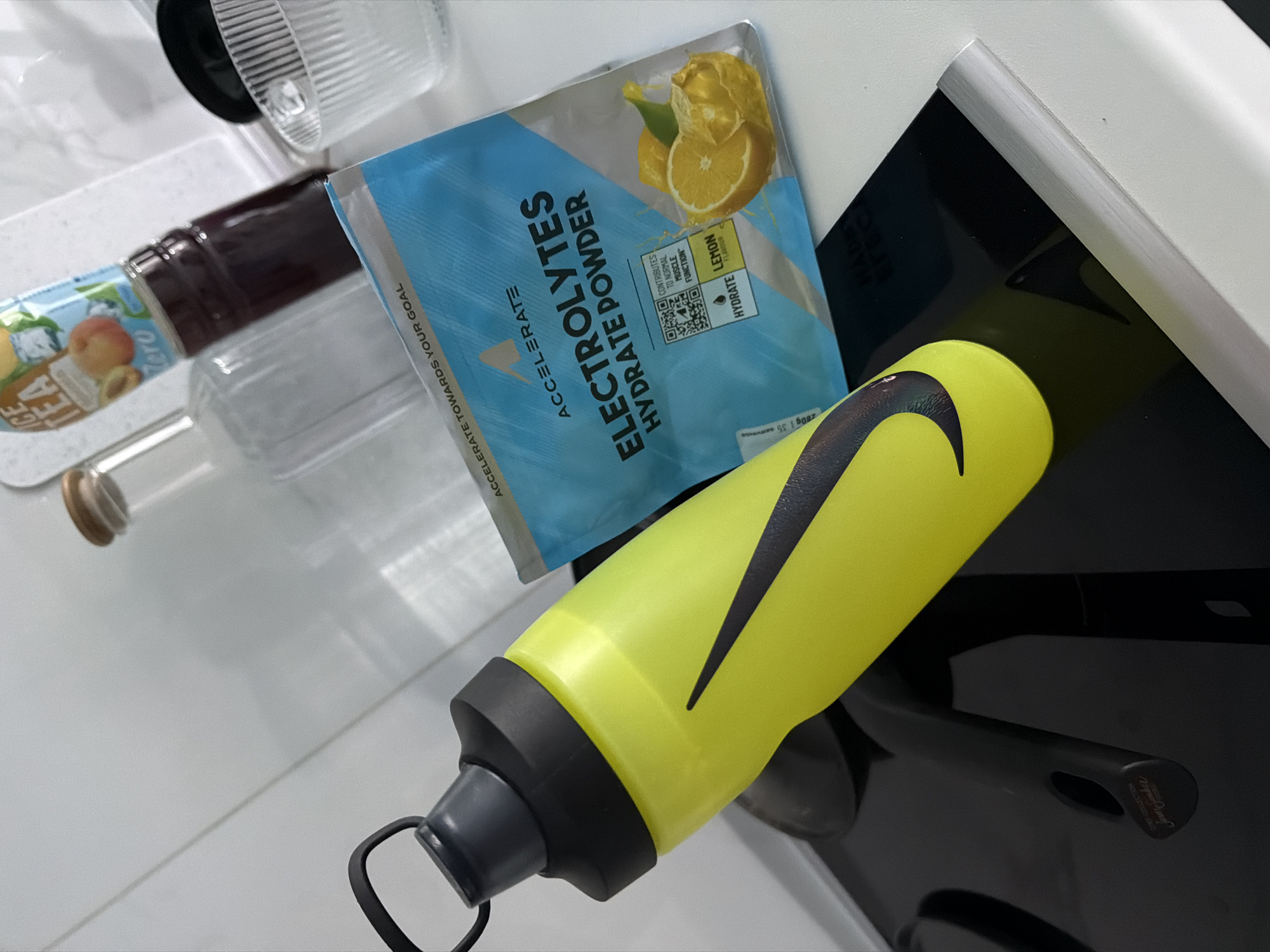 A bright yellow and black Nike water bottle is tilted on its side on a black surface, partially obscuring a package of "Accelerate Electrolytes Hydrate Powder" with a lemon design. Behind the powder package, a glass bottle containing a dark liquid and a clear glass bottle with a wooden stopper are visible. In the background, a ridged glass cup is also present. The scene is indoors, on a white tiled surface that appears to be a kitchen counter. The lighting is bright, suggesting an ambient light source. There is no indication of the city of Finestrat, Spain, in the image.