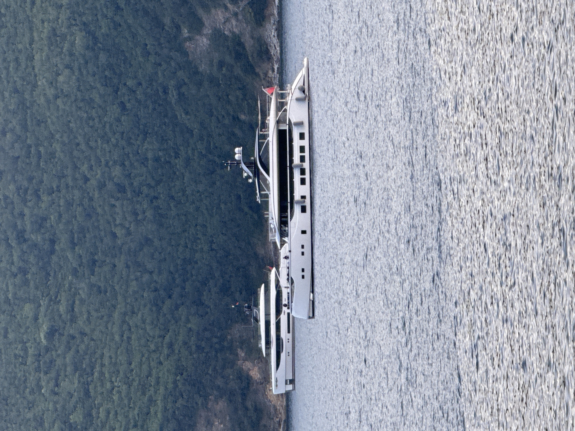 Two large, white motor yachts are moored in the calm, blue waters of the Adriatic Sea, with a densely forested green hillside forming the backdrop. The yacht in the foreground is closer to the viewer and positioned slightly to the right. It is a sleek, modern vessel with a dark-hulled lower section and white upper decks, featuring numerous rectangular windows along its sides. The yacht in the background is further away and to the left, also a white motor yacht with a similar contemporary design. Both vessels appear to be stationary. The environment suggests a tranquil, coastal setting, likely near a mountainous region. This scene is consistent with the coastal areas surrounding Budva, Montenegro, known for its picturesque bays and mountainous terrain.