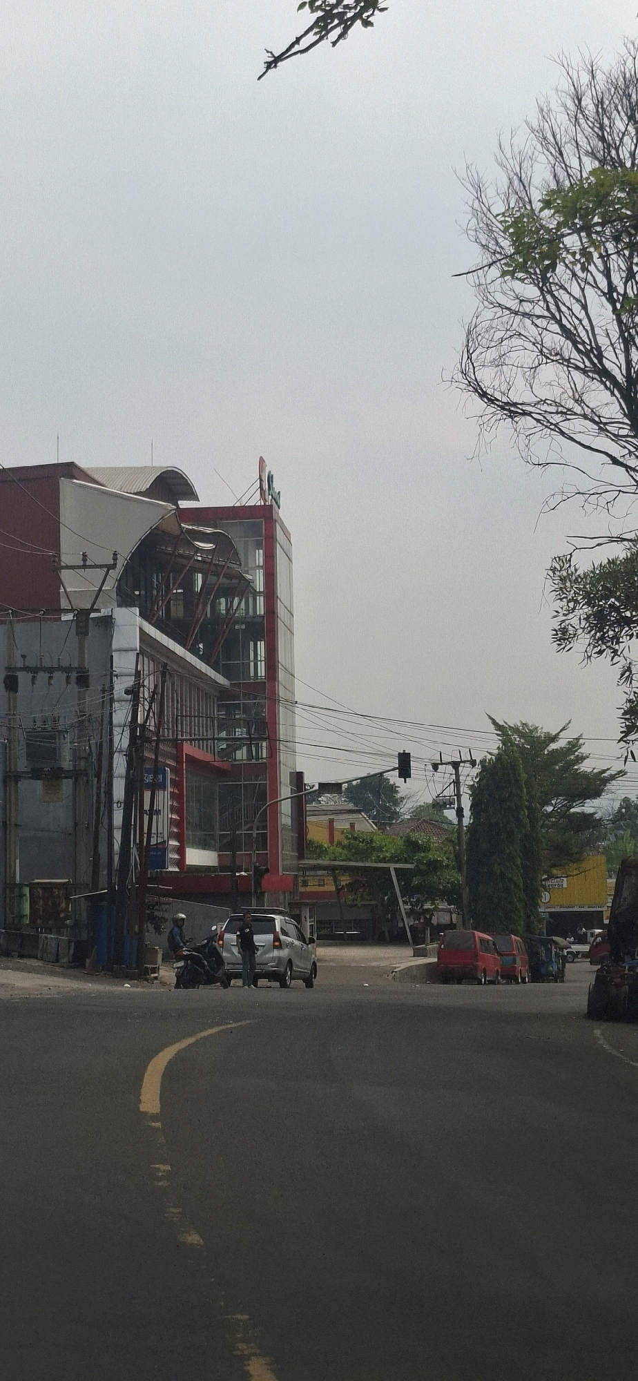 The image depicts a street scene in Sirnagalih, Indonesia, on an overcast day. In the foreground, a dark asphalt road features a yellow lane marking that curves from the bottom left towards the center. Several vehicles are present: a silver car is parked on the right side of the road, with a person standing next to it. To the left of the silver car, a motorcycle is parked, with a person in blue attire seated on it.
Dominating the left side of the frame is a multi-story building with a red and gray facade. The building has large windows and a distinctive curved roof structure. Electrical poles and wires are visible in front of this building. Across the street, a row of smaller buildings with yellow and red accents can be seen. Further in the background, lush green trees and foliage line the horizon. In the middle ground, to the right, two red vans are parked side-by-side. The overall atmosphere is one of a typical urban environment, with clear signs of daily activity.