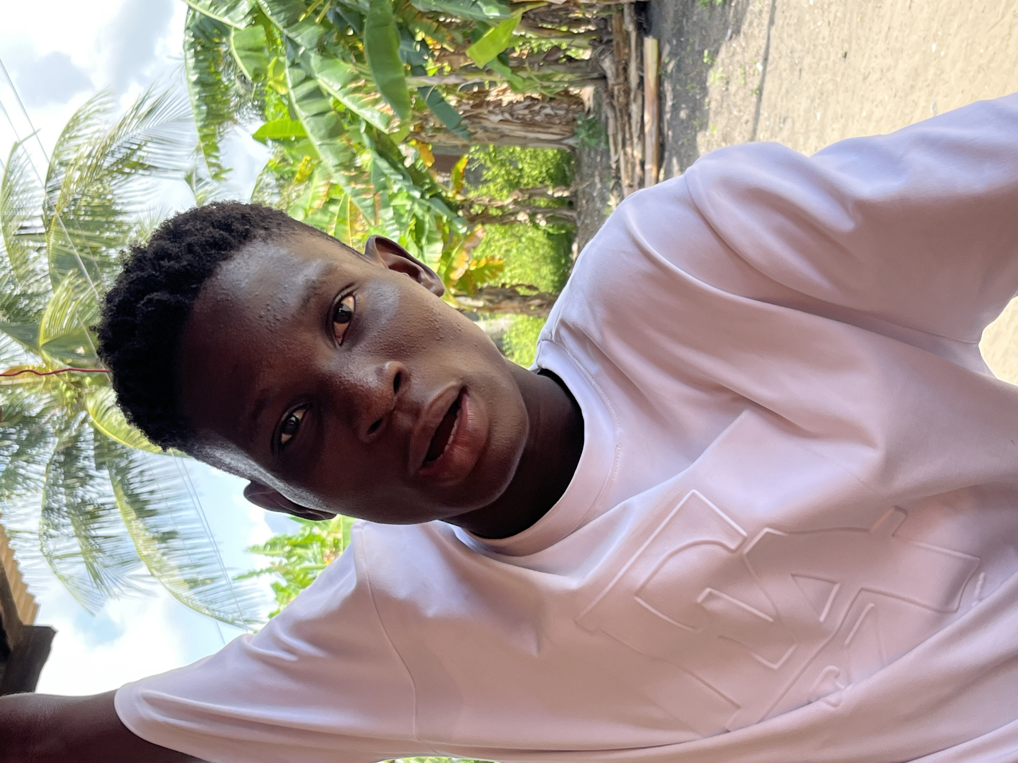 A close-up angled outdoor shot depicts a young dark-skinned African male wearing a white long-sleeved shirt with an embossed design on the front. The subject is turned slightly to his right, looking toward the camera with a somewhat open mouth. The background features tropical foliage, including banana trees and what appears to be a palm tree, hinting at a warm, humid climate. This image was taken in Badagry, Nigeria.