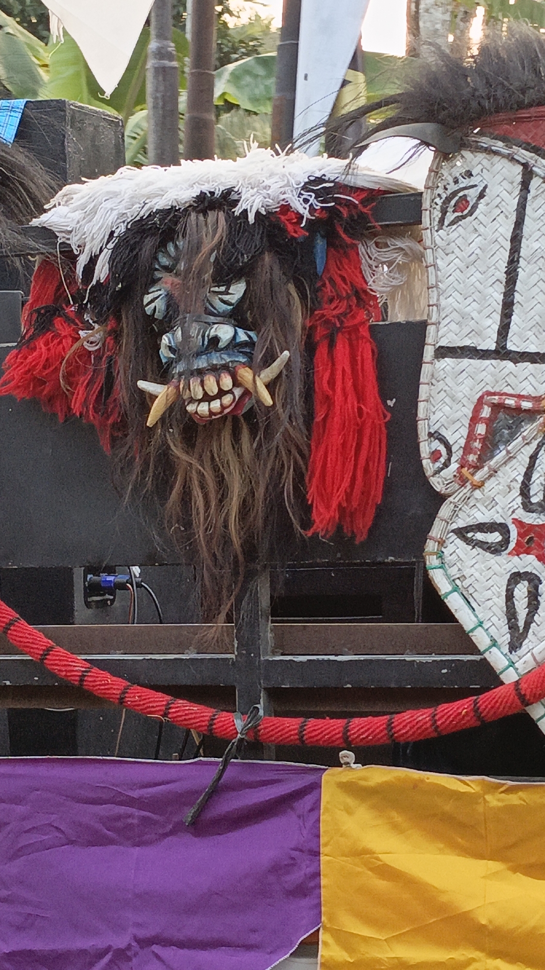 In Krajan Lor, Indonesia, a close-up shot showcases a decorative, menacing mask mounted on a dark, textured surface, possibly a stage or backdrop. The mask features a fearsome face with bulging, light-blue eyes, a wide mouth with sharp, yellow tusks protruding, and a reddish-pink tongue. It is adorned with a wild mane of dark, brown, and black hair, complemented by vibrant red and white fringe. To the right of this mask, a portion of another mask, crafted from woven material with a white background, is visible. This second mask has a stylized red eye and intricate black patterns. A thick, red rope with black accents is strung across the lower part of the image, tied with a dark cord. Below the rope, swathes of fabric in bright purple and yellow are draped, suggesting a festive or ceremonial setting. The background consists of greenery, possibly foliage or banana leaves, and thin, vertical poles, indicating an outdoor location.